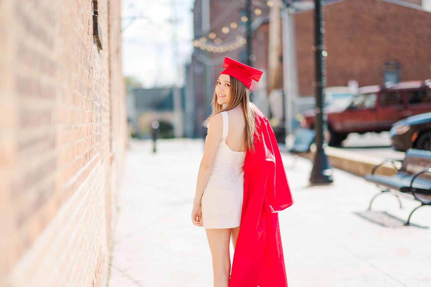 Graduate in red cap and gown walks along a brick-lined urban sidewalk, glancing back.