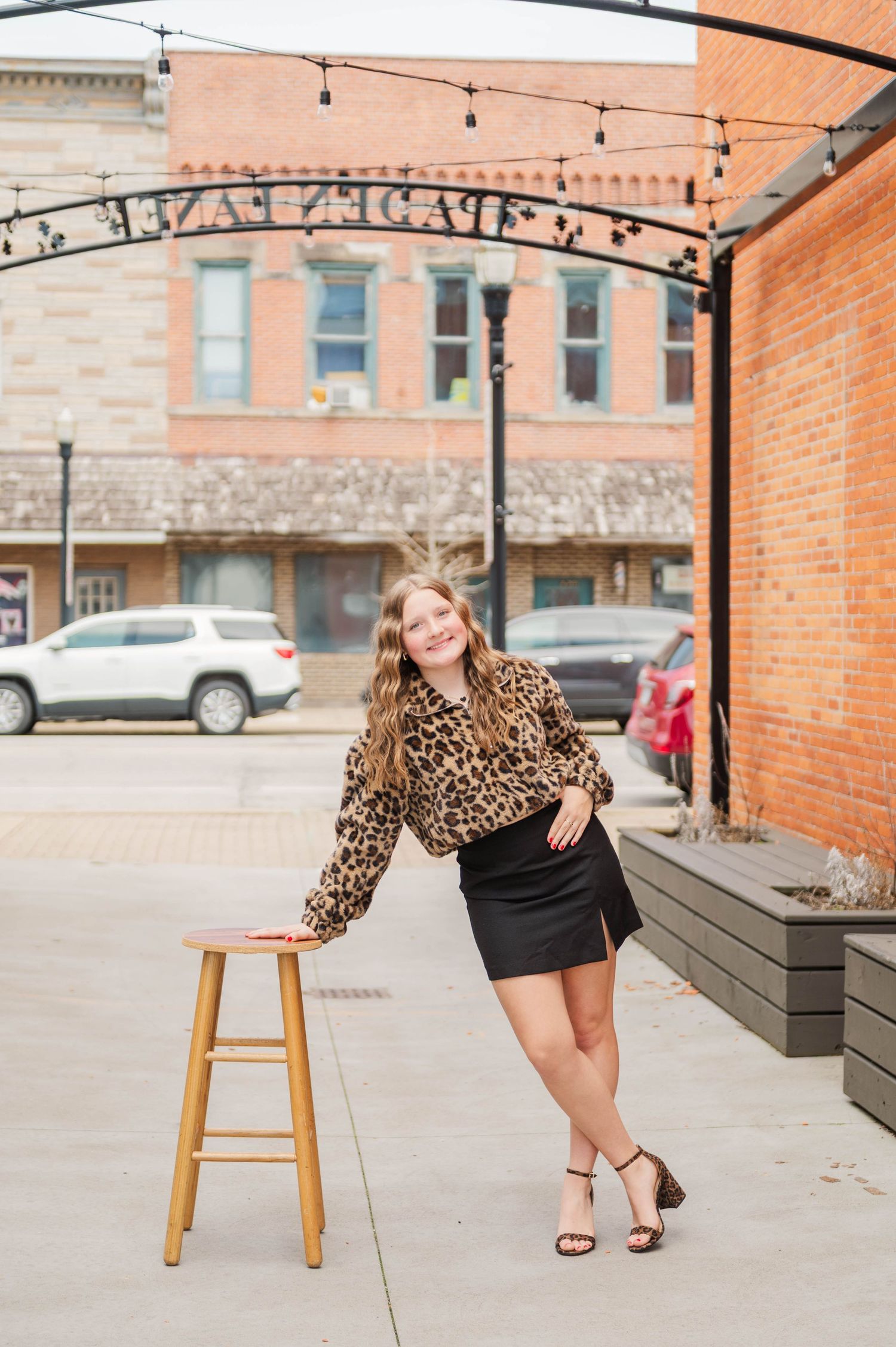 Young woman in leopard print top and black mini skirt posing by a wooden stool in a small town outdoor setting.