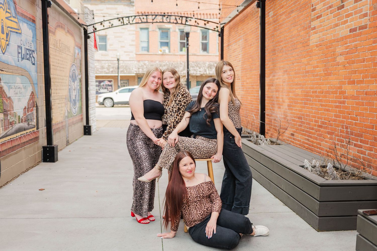 Five young women pose together in animal print outfits along brick-lined outdoor corridor with string lights.