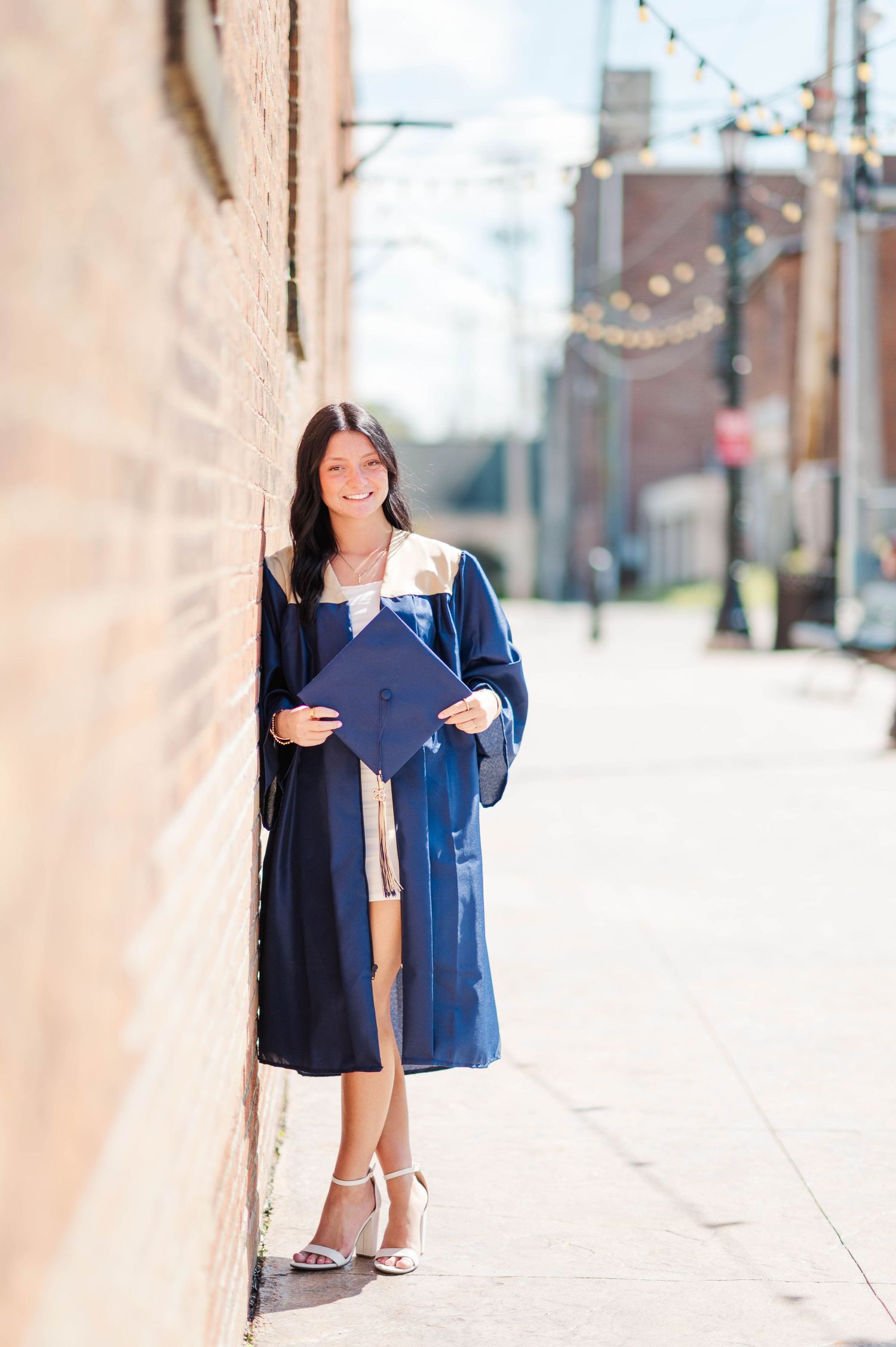 Graduate in navy blue cap and gown leaning against brick wall on urban sidewalk.
