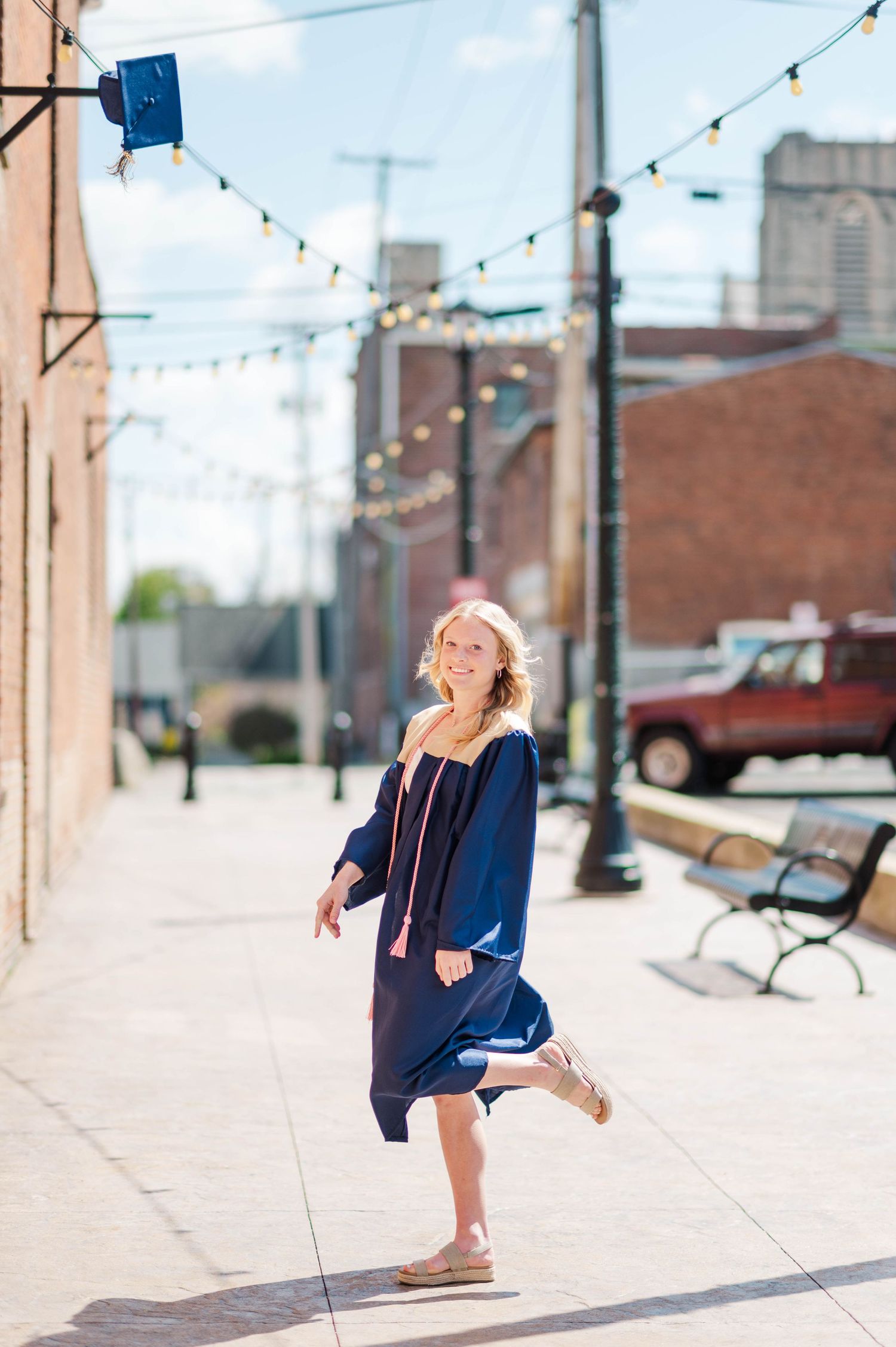 Blonde graduate in navy gown tossing cap while dancing on sunny sidewalk with string lights.
