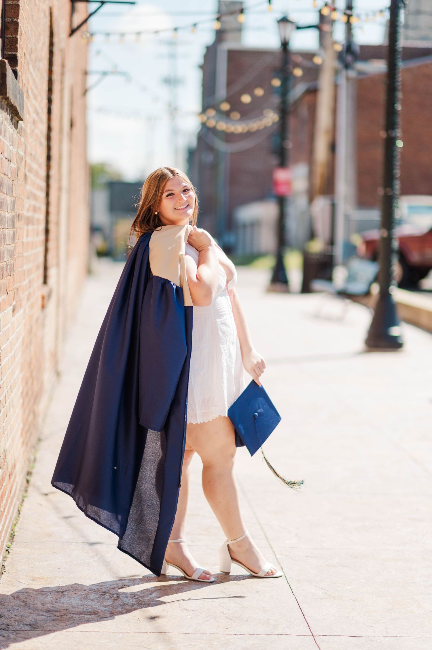 Cheerful graduate in white dress and navy gown walks along sunny brick alley holding mortarboard cap.