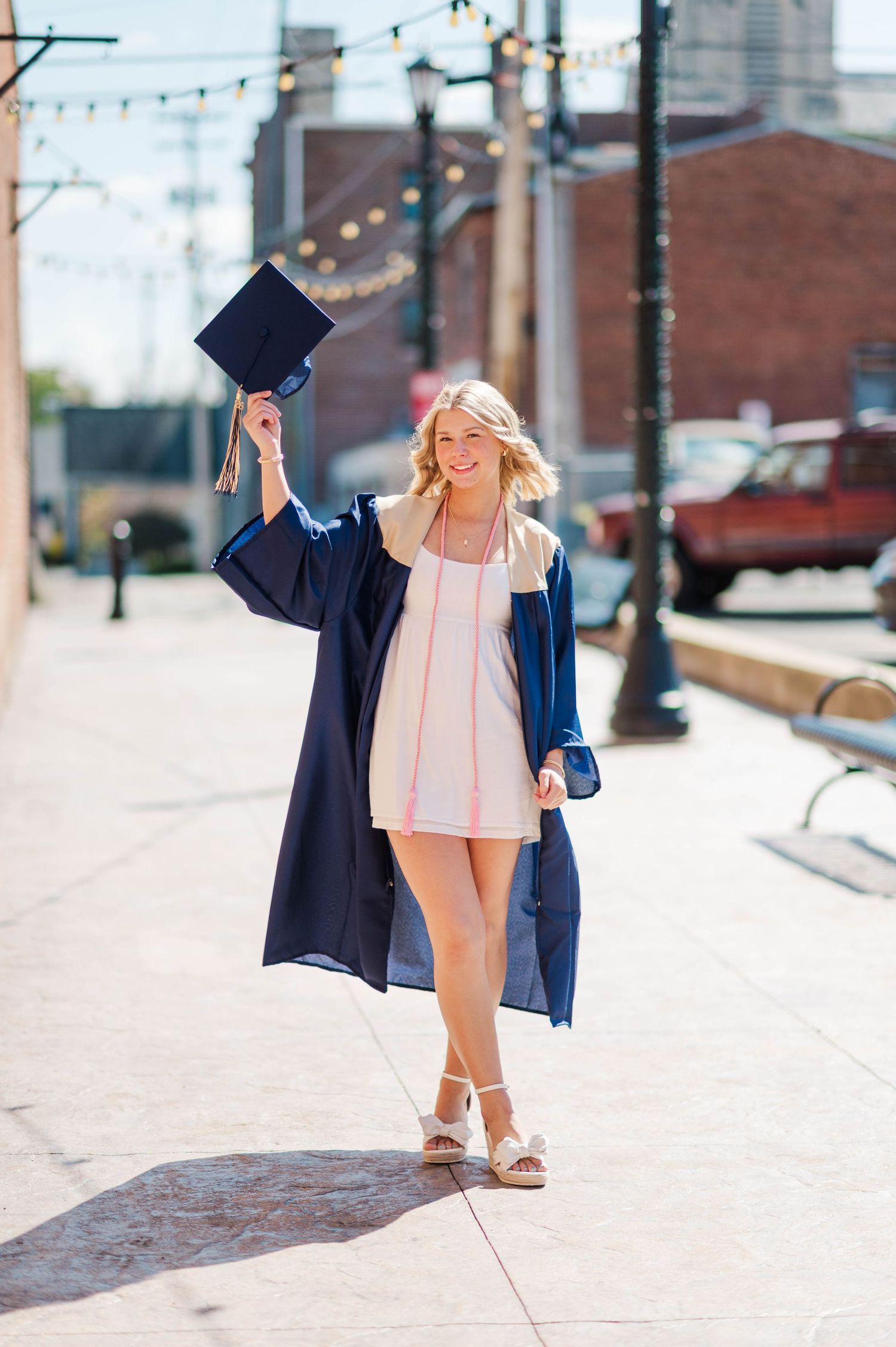 Smiling graduate in navy cap and gown holding cap up on sunny urban sidewalk.