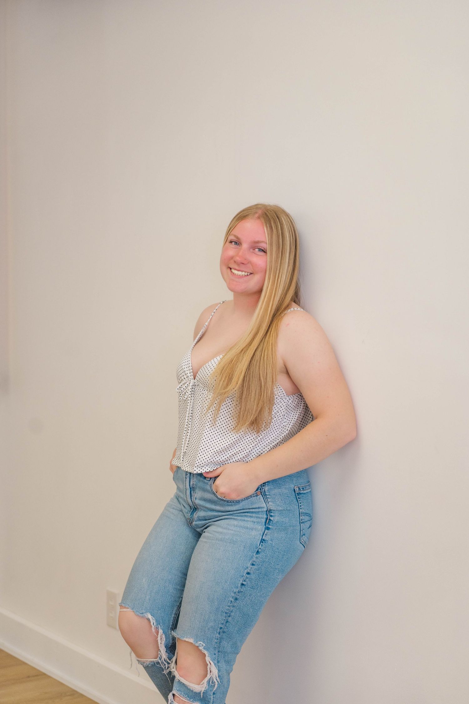 Smiling blonde woman in striped top and ripped jeans leaning against a white wall.
