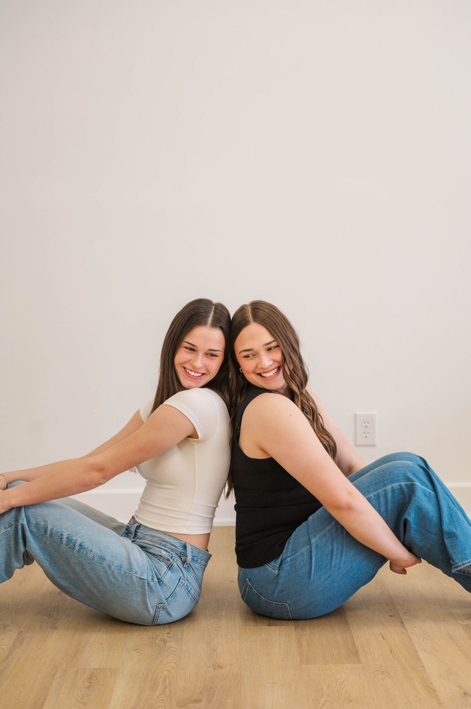 Two smiling young women in jeans sitting back-to-back on hardwood floor against white wall.