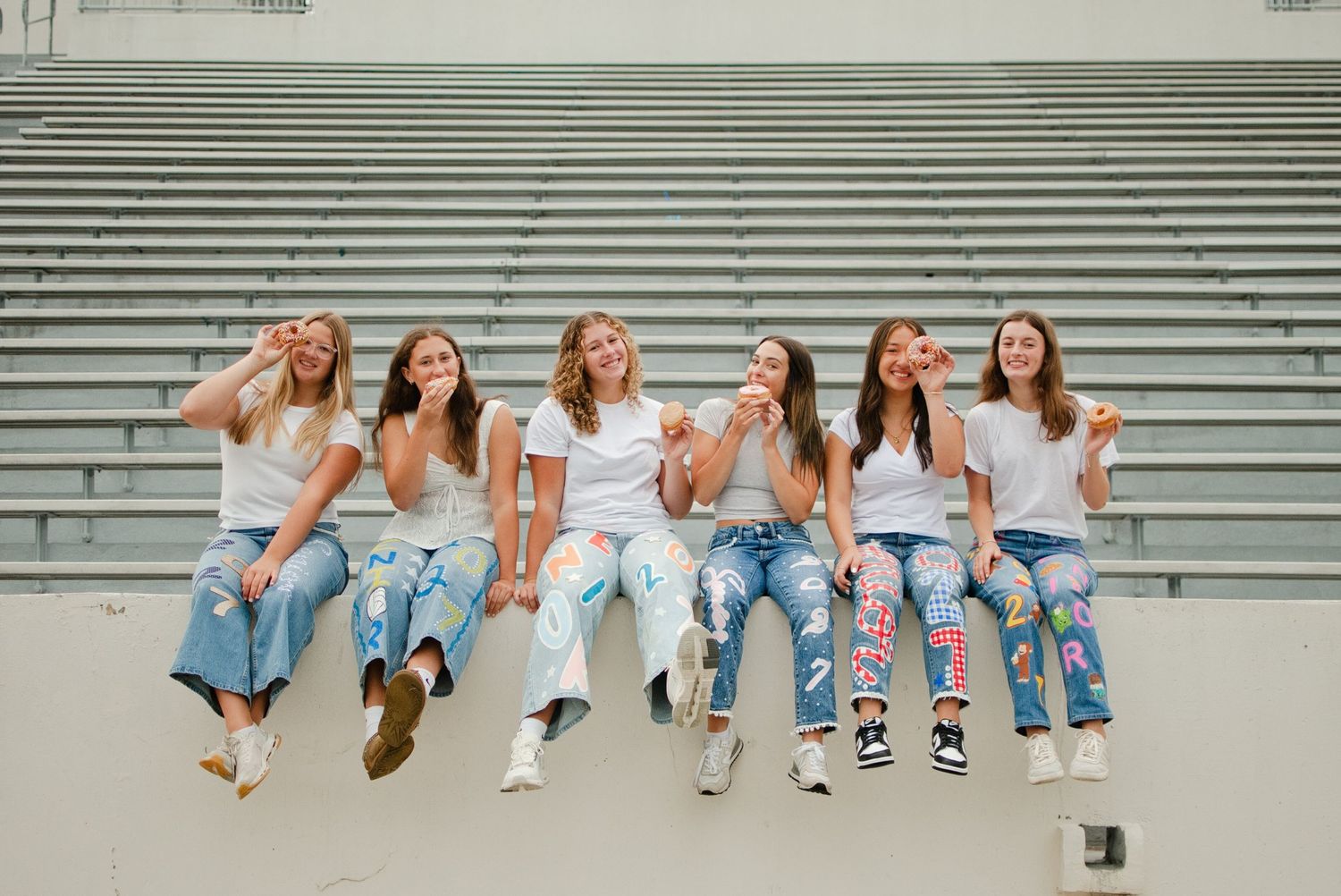 Six teenage girls in white shirts and decorated senior jeans sit laughing together on stadium bleacher steps.