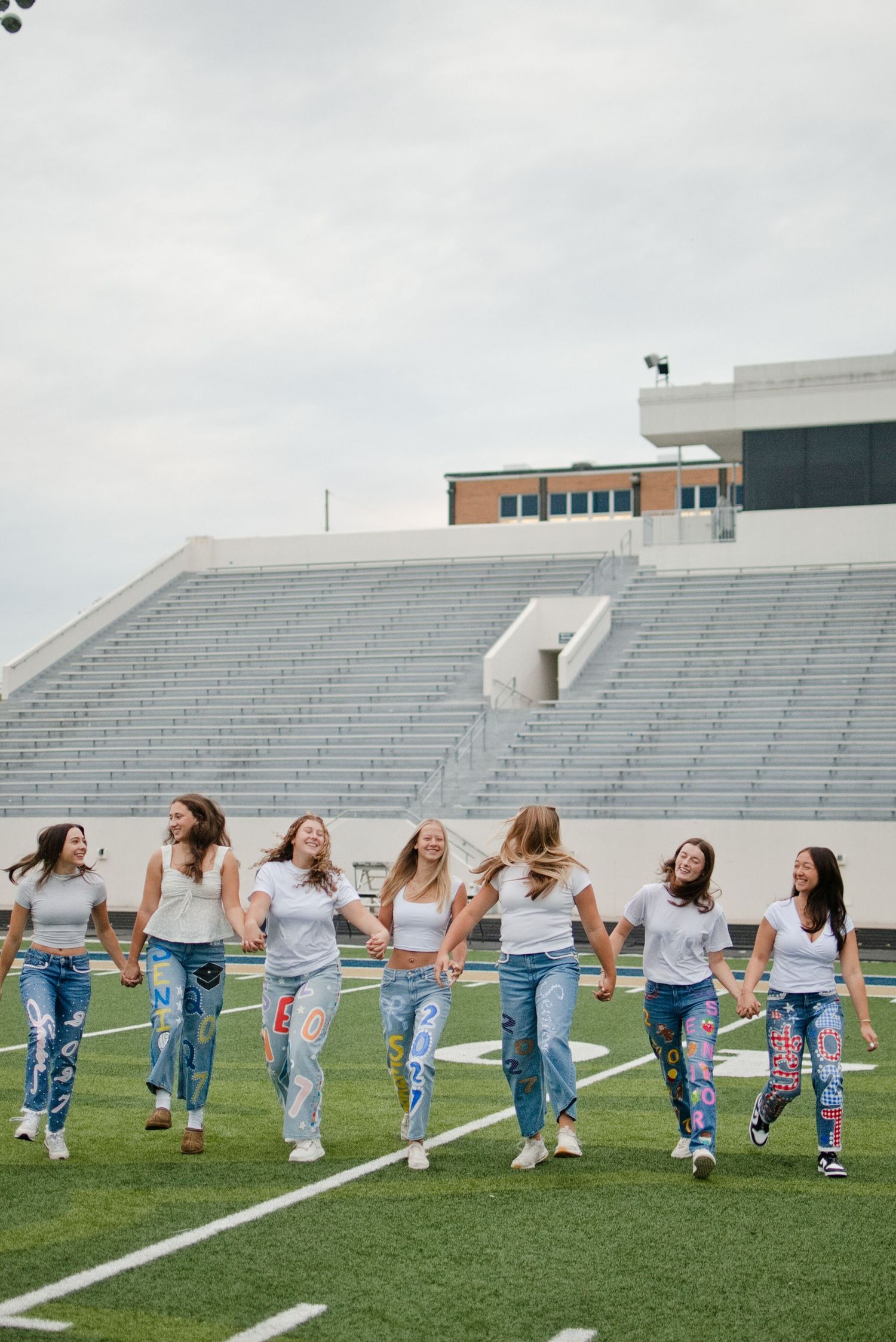 Group of seven teenage girls in matching white shirts and custom senior jeans walking and laughing on a football field.