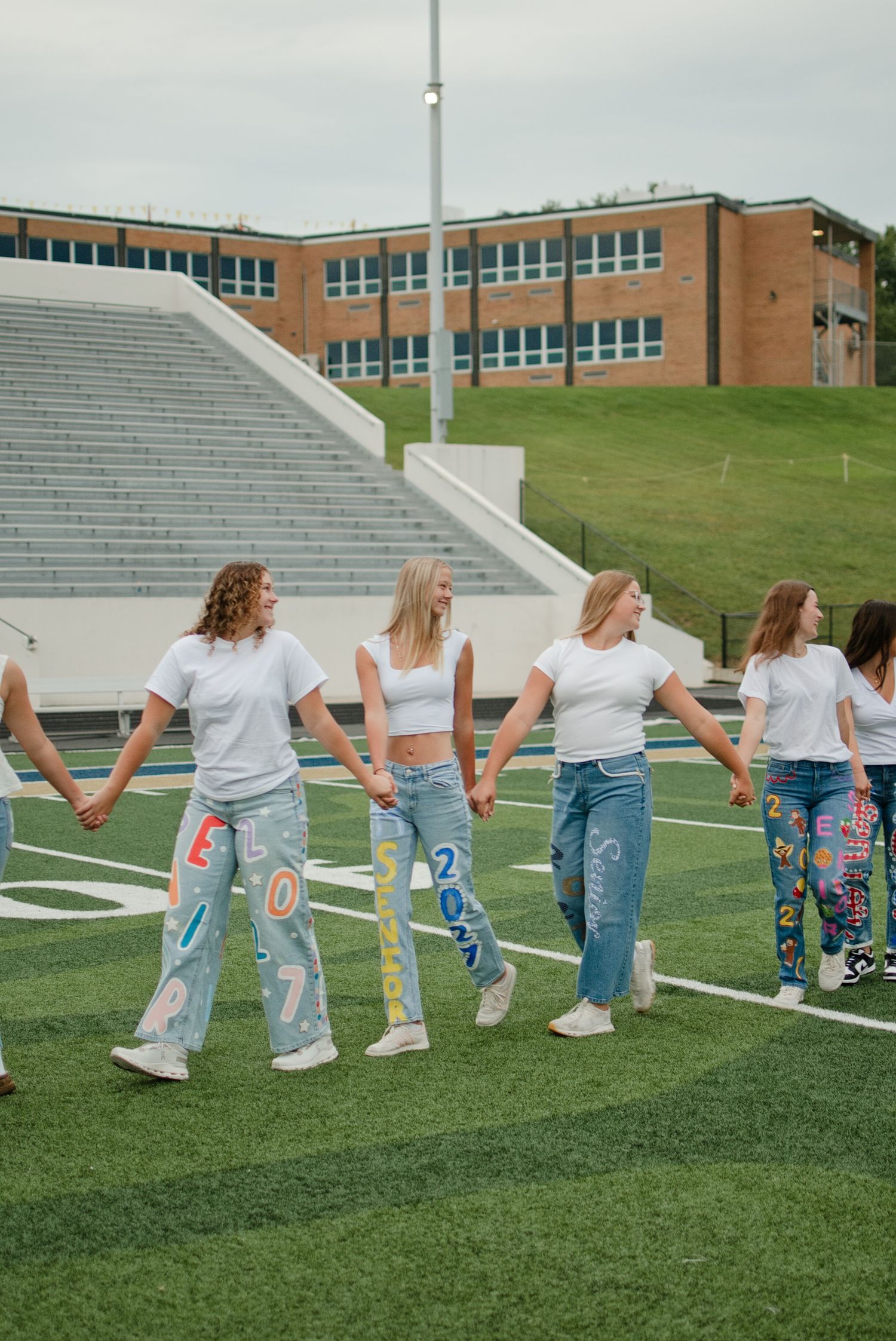 Group of teen girls in white tops and custom painted jeans holding hands on a high school football field.