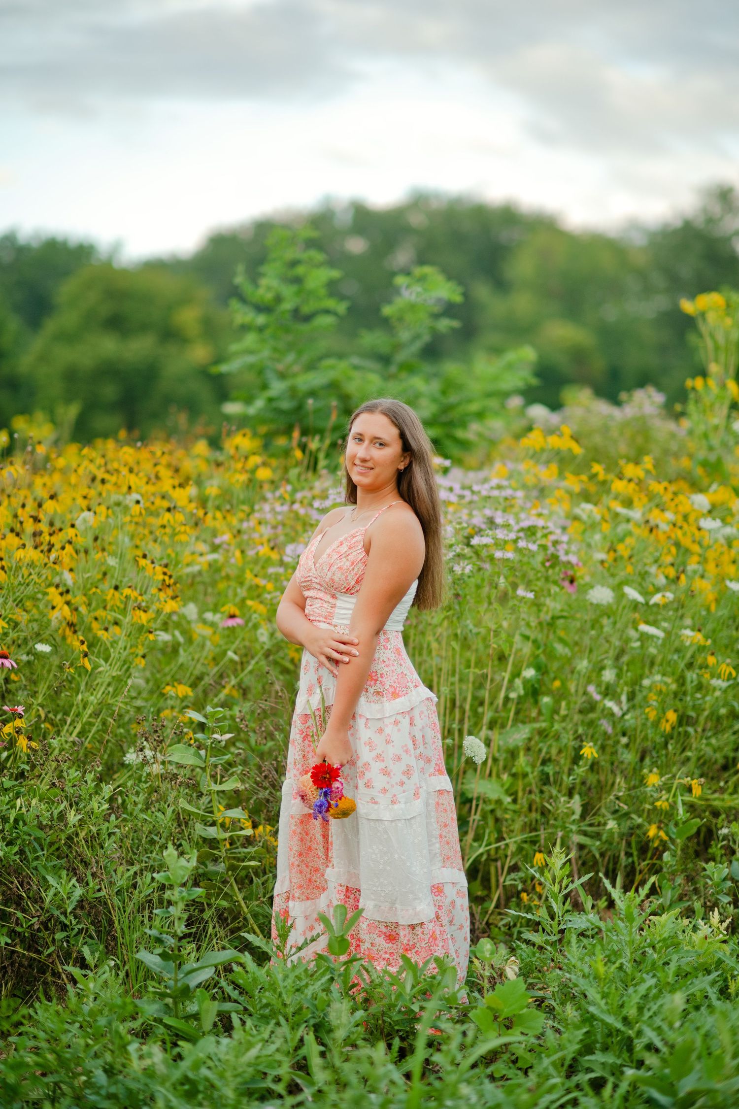 Young woman in floral patchwork maxi dress standing in vibrant wildflower meadow holding colorful blooms.