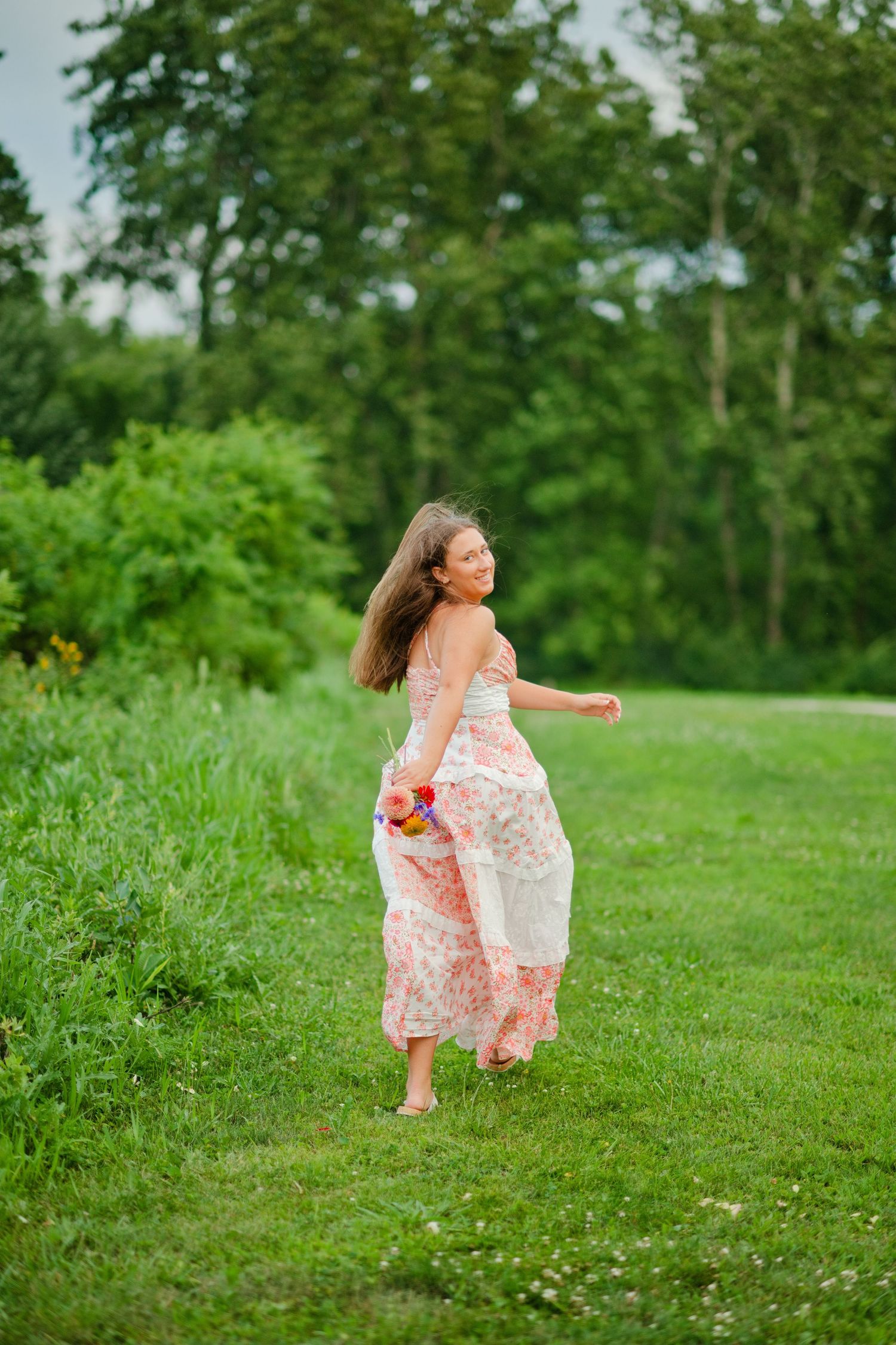 Young woman in a pink floral maxi dress twirling barefoot on a lush green lawn surrounded by trees.