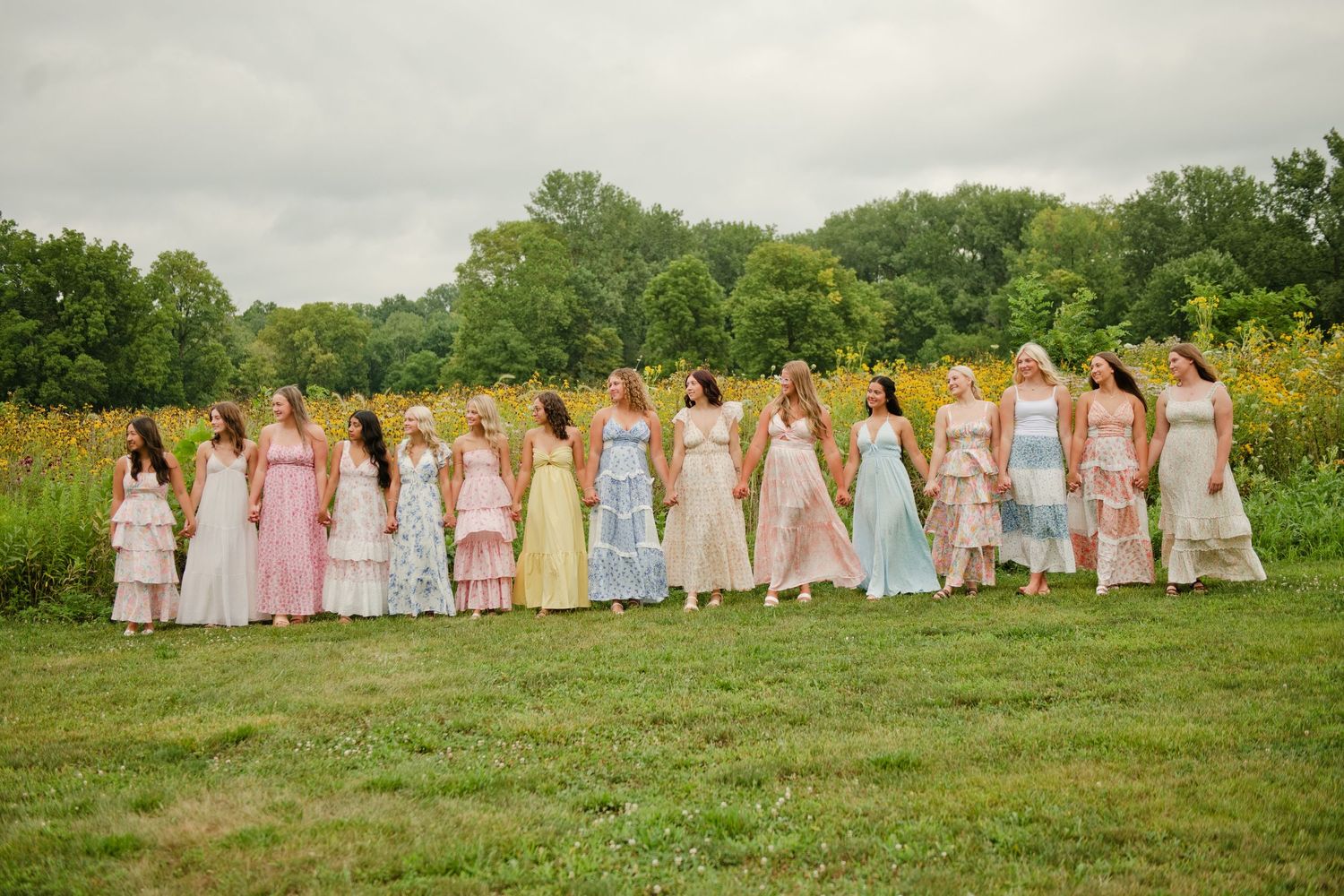 A line of bridesmaids in colorful mismatched floral dresses standing in a green meadow on a cloudy day.