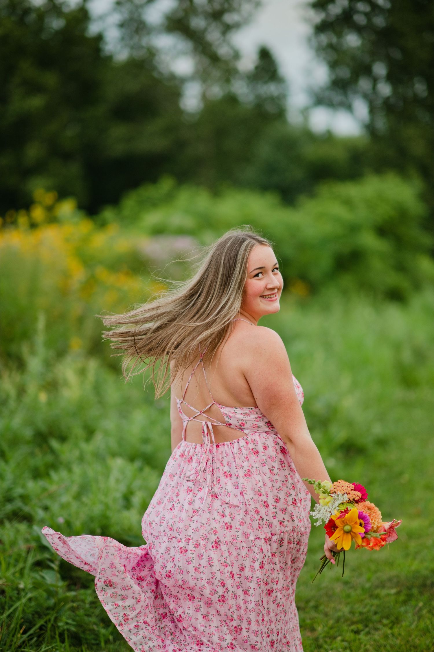 Smiling blonde woman in a pink floral dress holding a colorful bouquet while twirling in a green meadow.