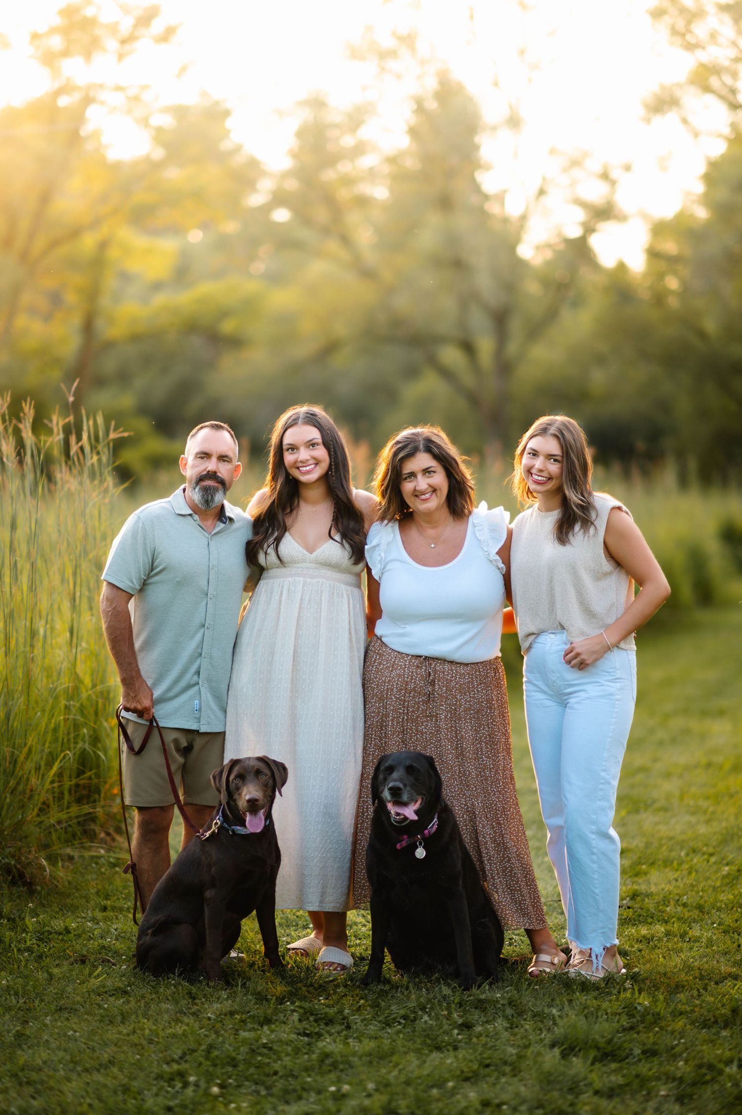 A family poses with their two black dogs in a sunlit meadow during golden hour.