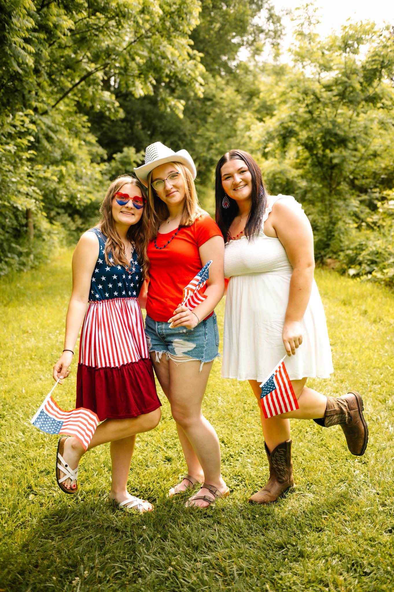 Friends wearing patriotic red white and blue outfits pose playfully in a sunny meadow with American flags.