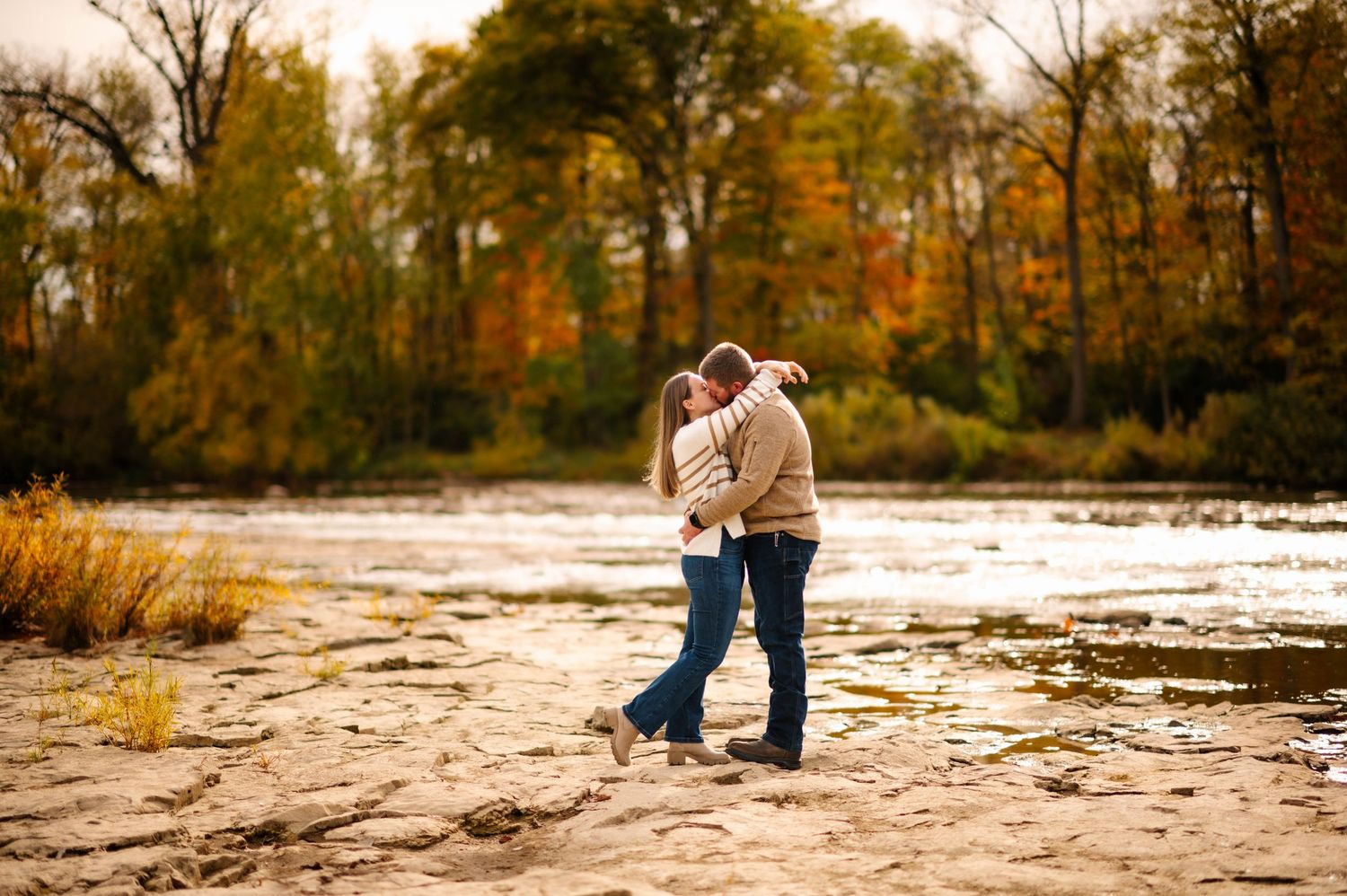 Couple poses together on rocky riverbed with autumn foliage in background.