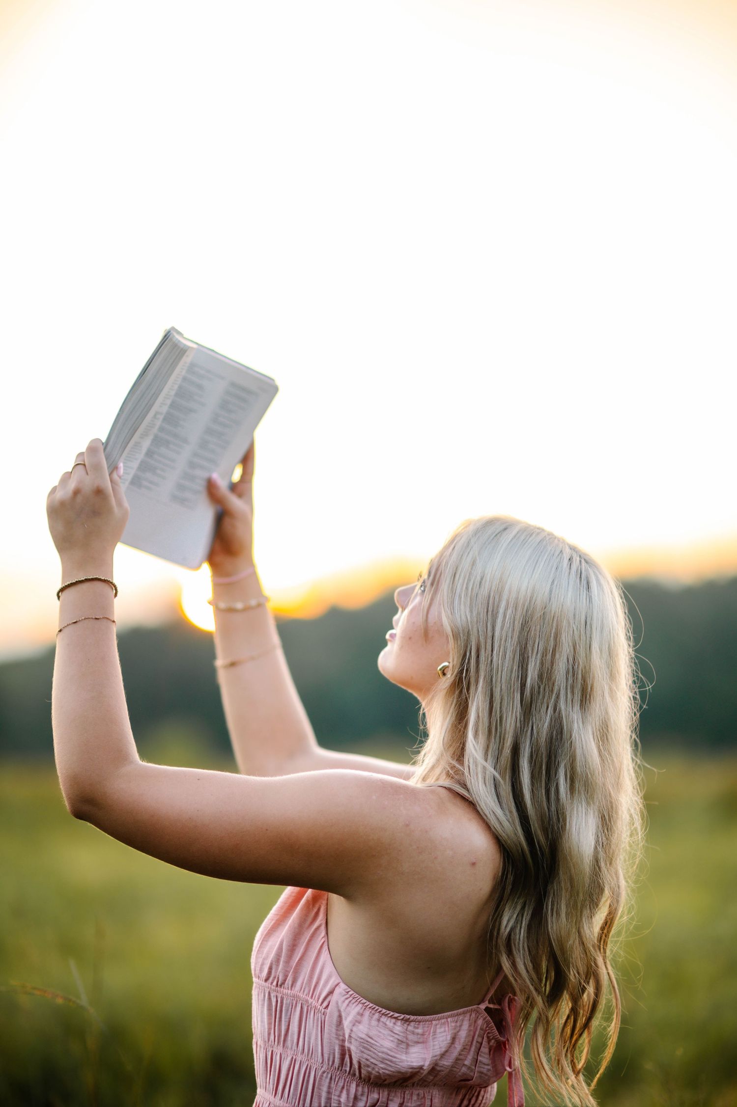 Someone in a pink sundress holds up papers against a glowing sunset sky in a meadow.