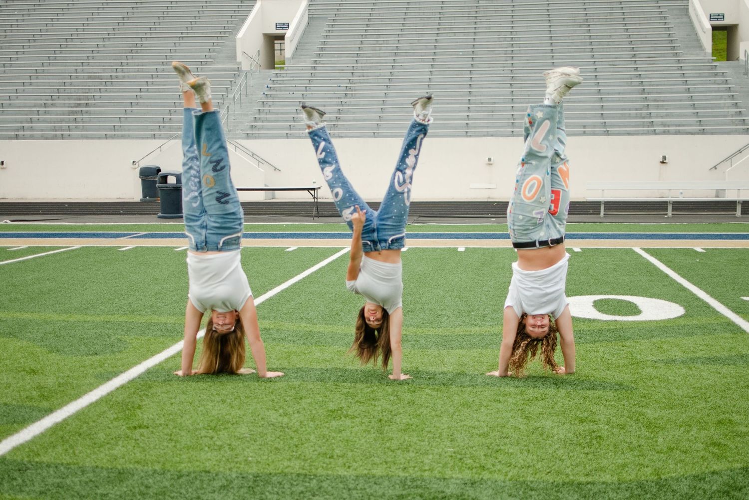 Three girls doing handstands on a football field turf wearing jeans and white tops.