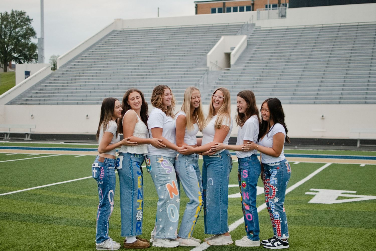 Seven young women in white tops and jeans pose together on a football field stadium turf.