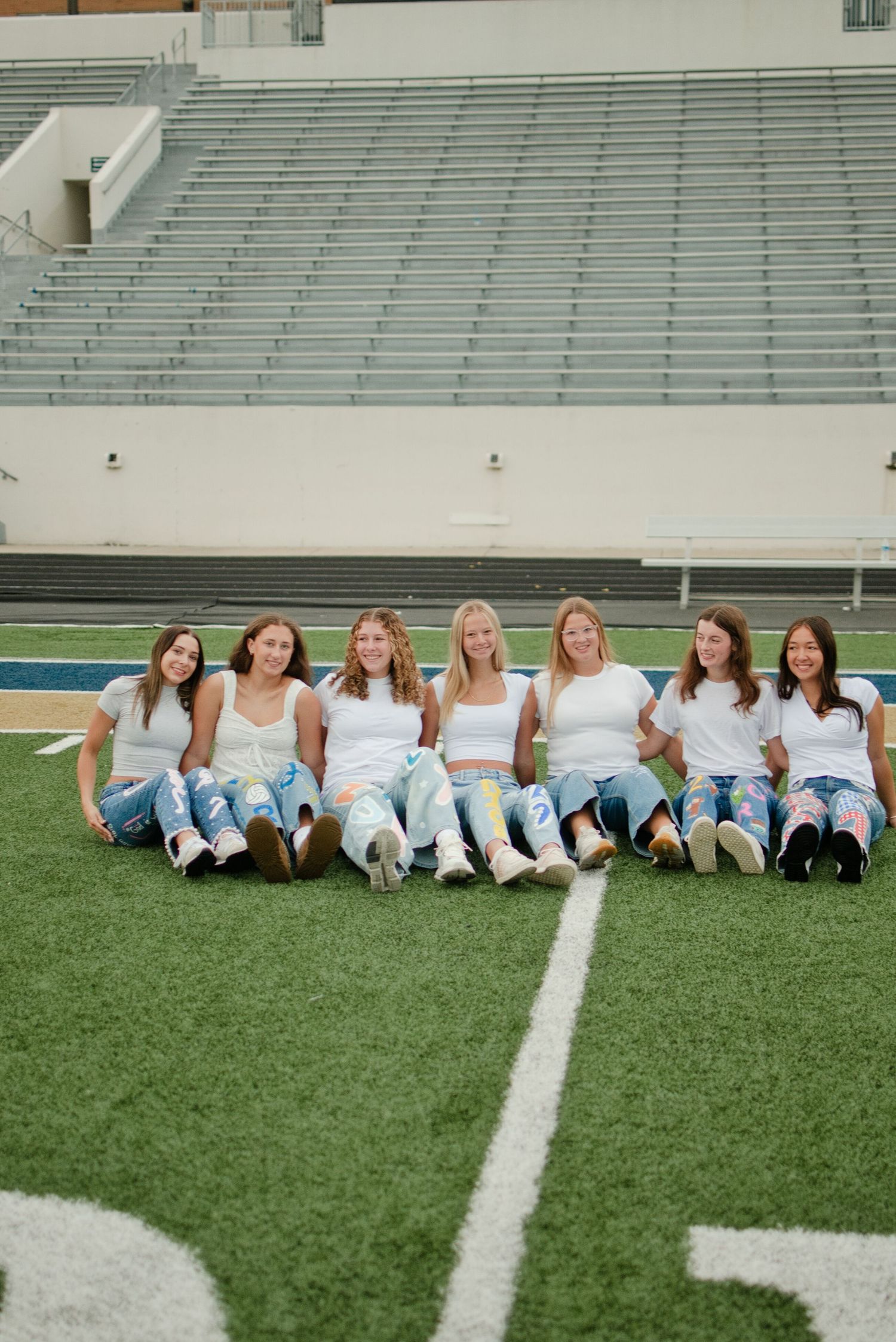 Seven smiling teen girls in white tops and custom senior jeans sit in a row on a football field turf.