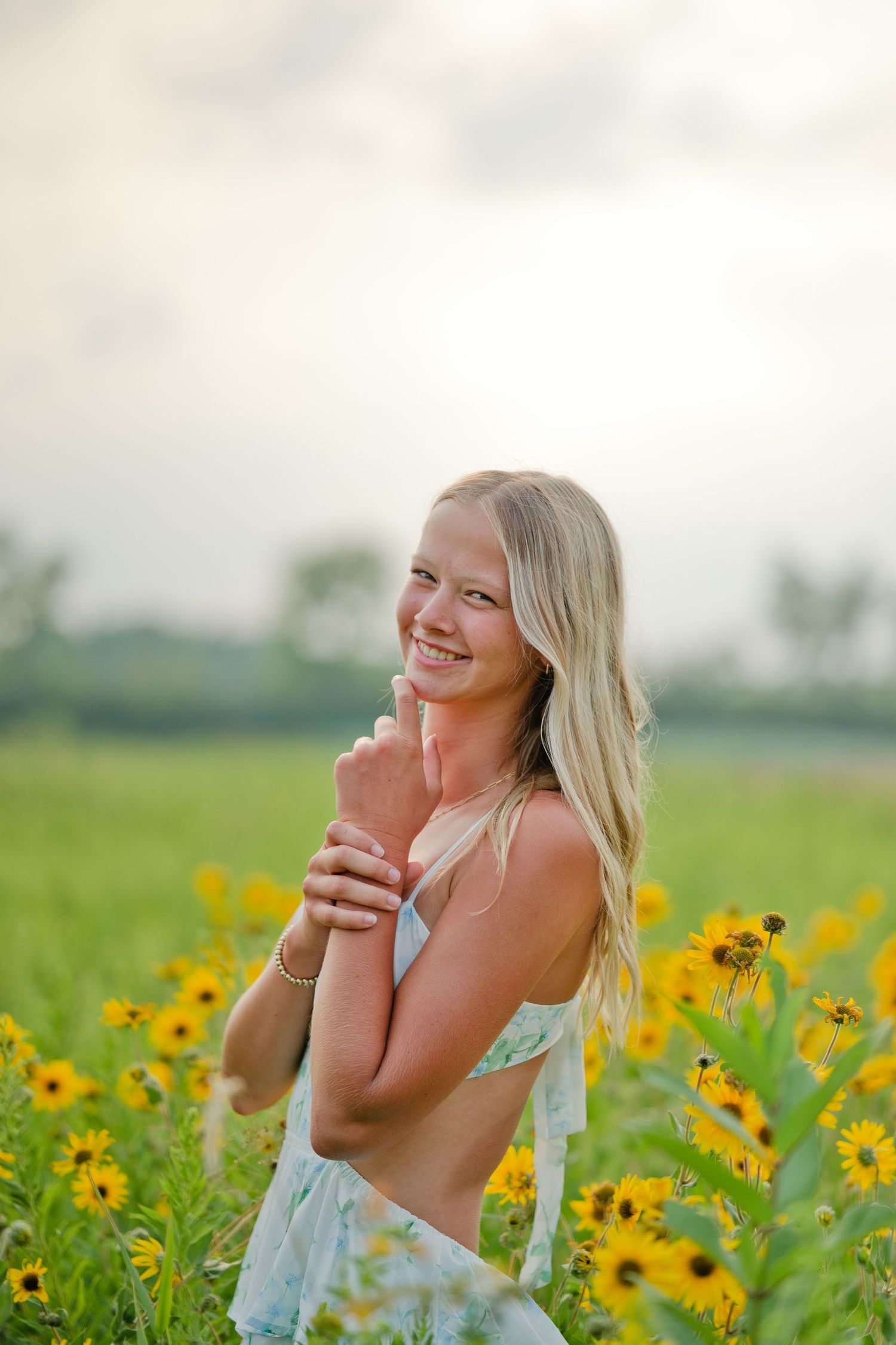 Smiling blonde woman in floral dress standing among yellow wildflowers in a sunny green field.