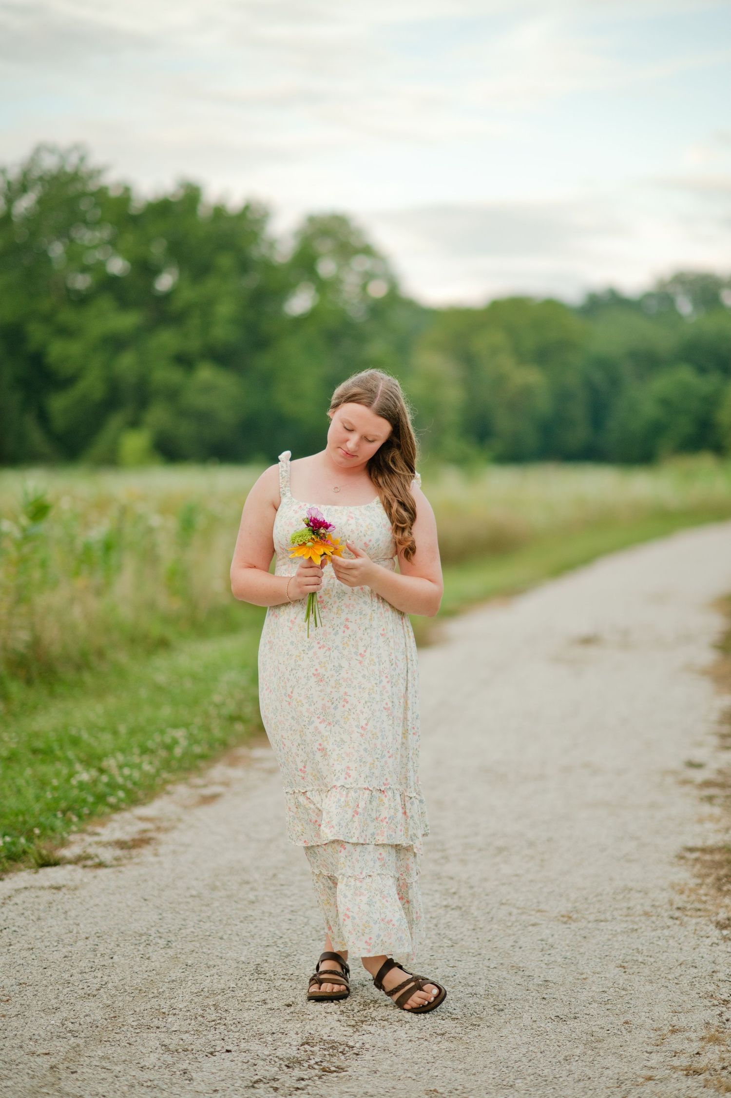 Woman in white floral dress holding a small bouquet on a gravel country path.
