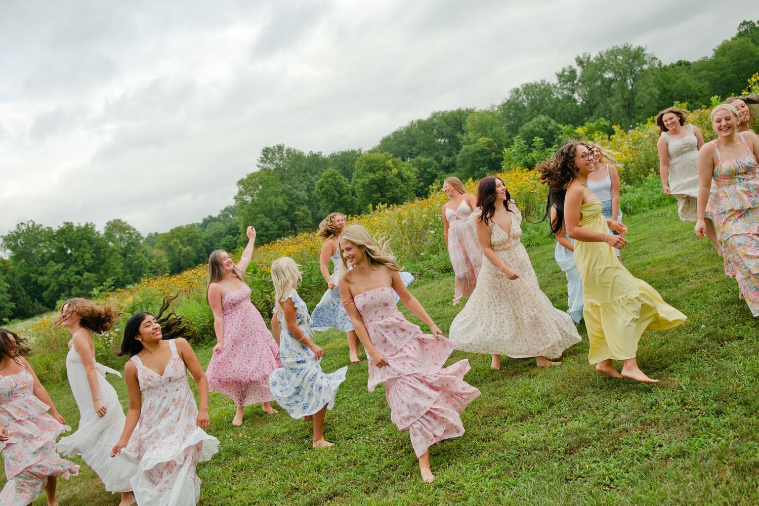 Group of women in floral pastel dresses running joyfully on a green hillside on a cloudy day.