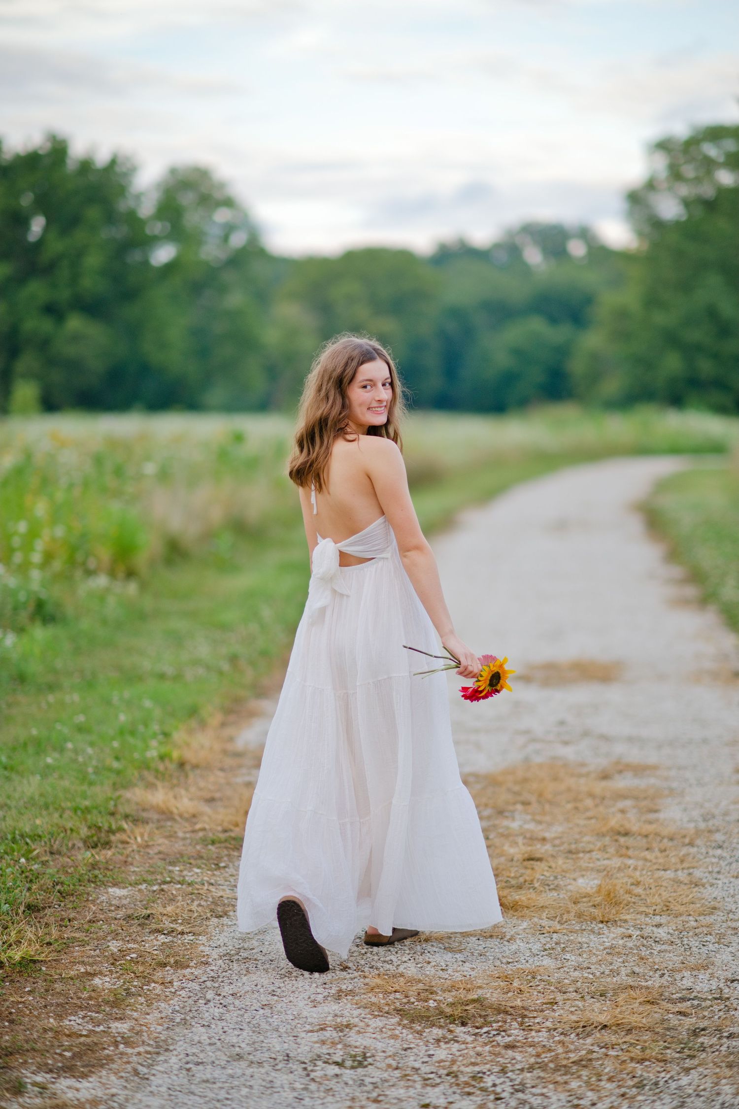 Young woman in white backless gown holding flowers on a scenic gravel path.