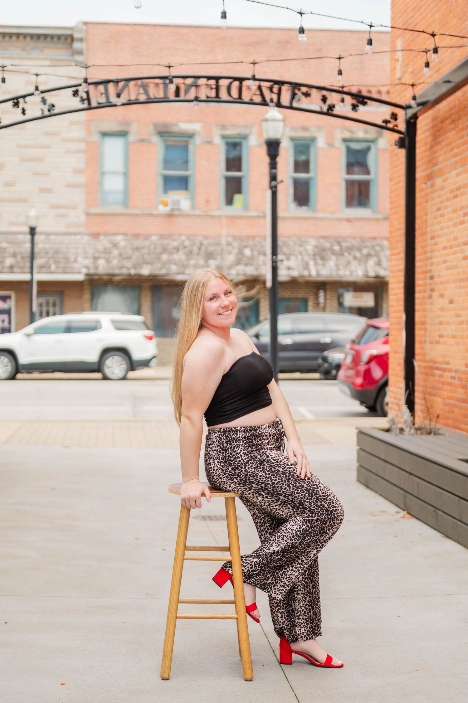 Young blonde woman poses on stool in leopard pants and black crop top with red heels in downtown setting.