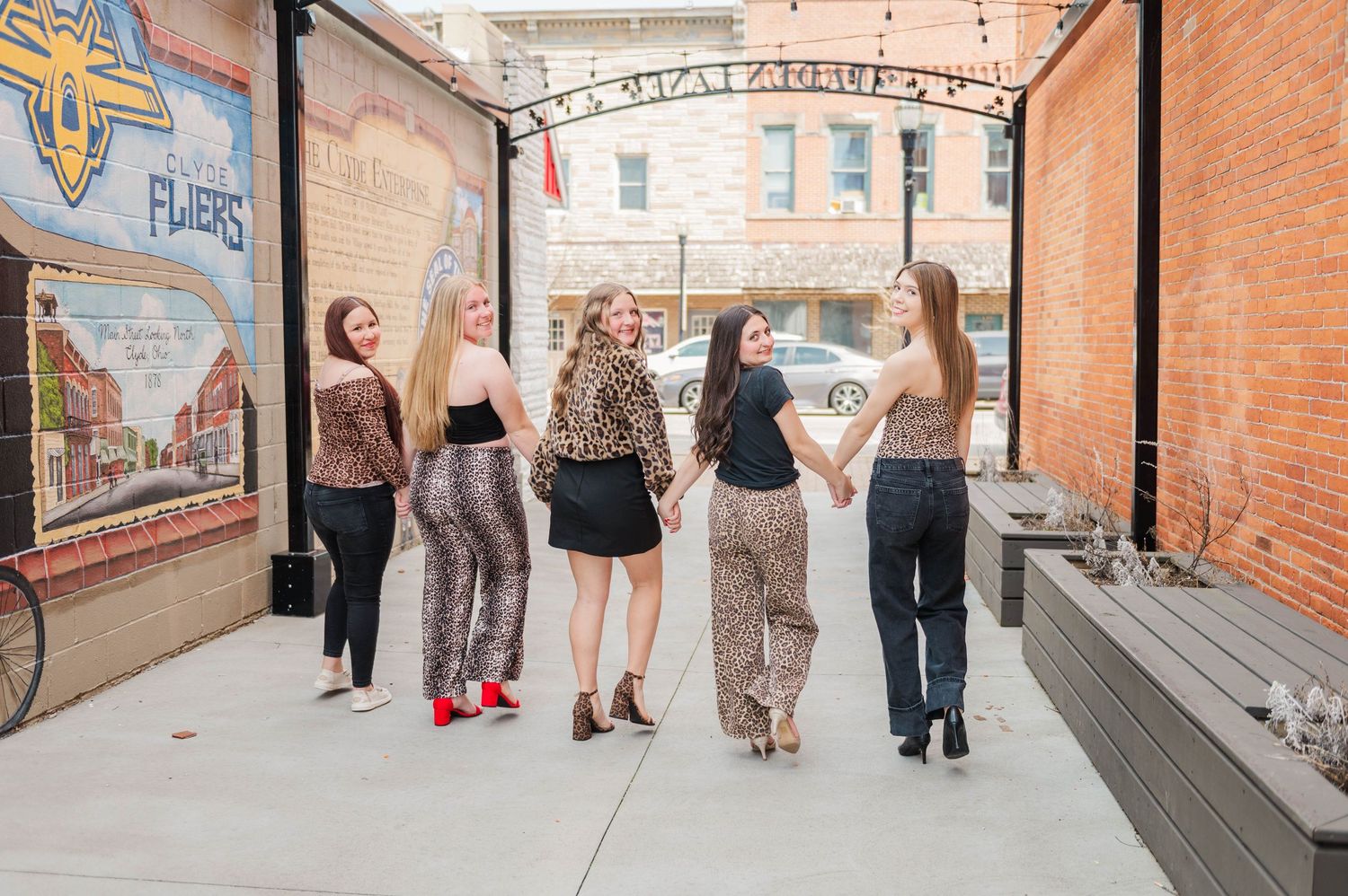 Five stylish young women in leopard print clothing walk together holding hands on an outdoor patio.