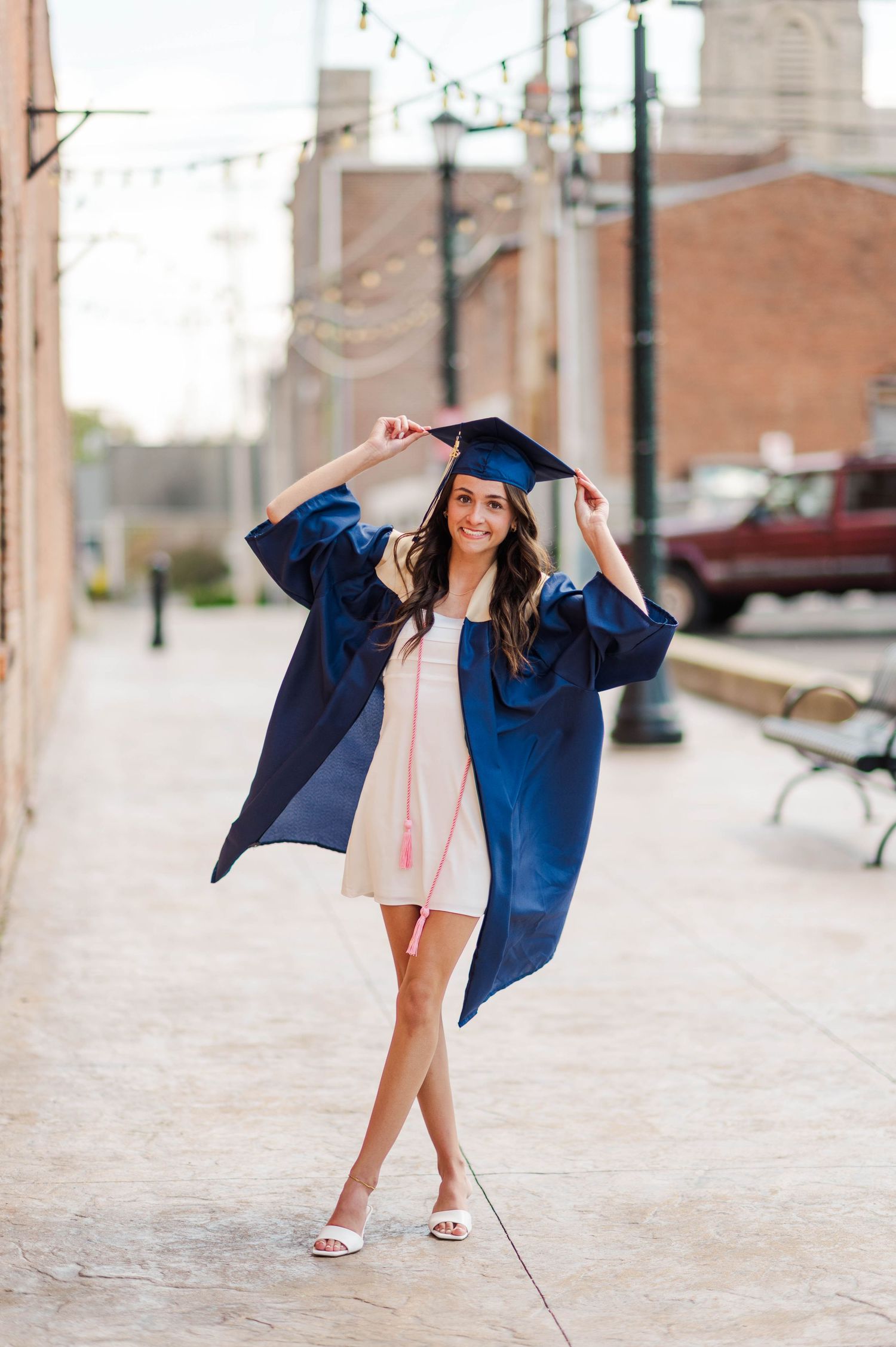Smiling graduate in navy cap and gown over white dress posing outdoors on brick sidewalk.