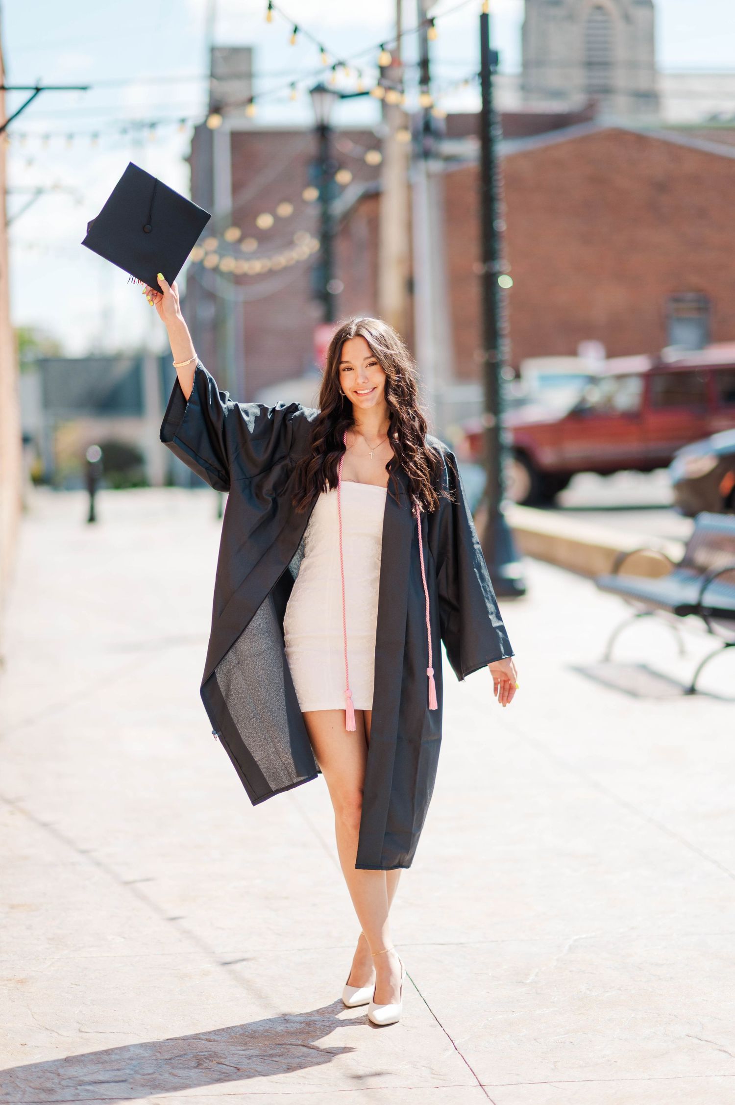 Brunette woman in black graduation gown holding cap up while walking on sunny urban sidewalk.