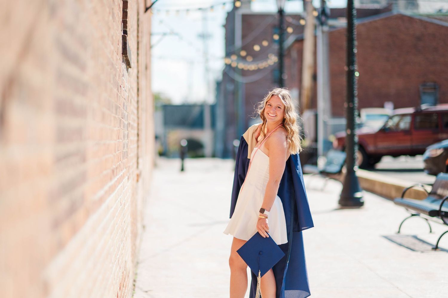 Young woman in navy graduation gown walks along sunny brick-lined urban alley.