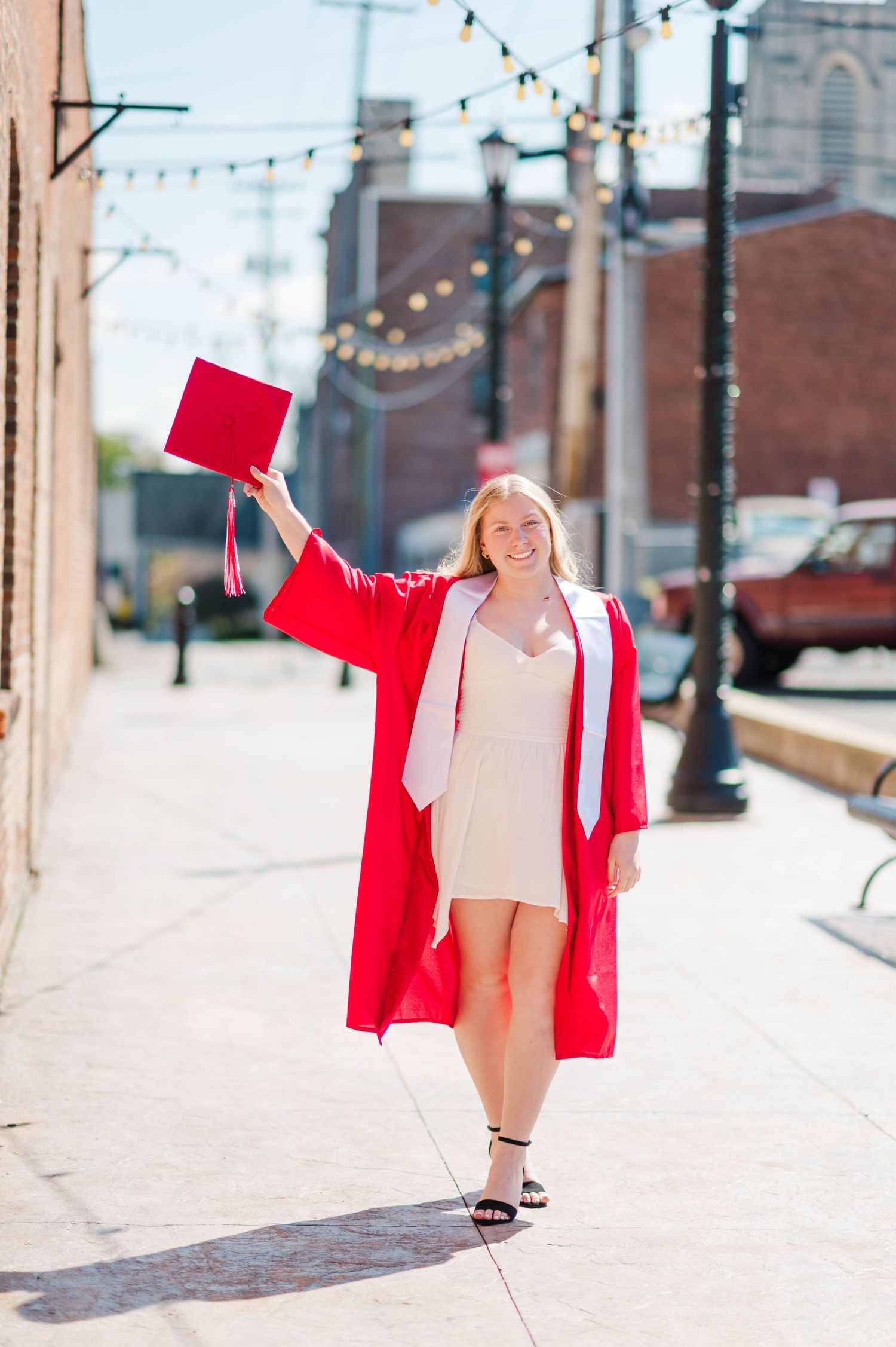 Smiling graduate in red cap and gown raises her cap triumphantly on a sunny urban sidewalk.