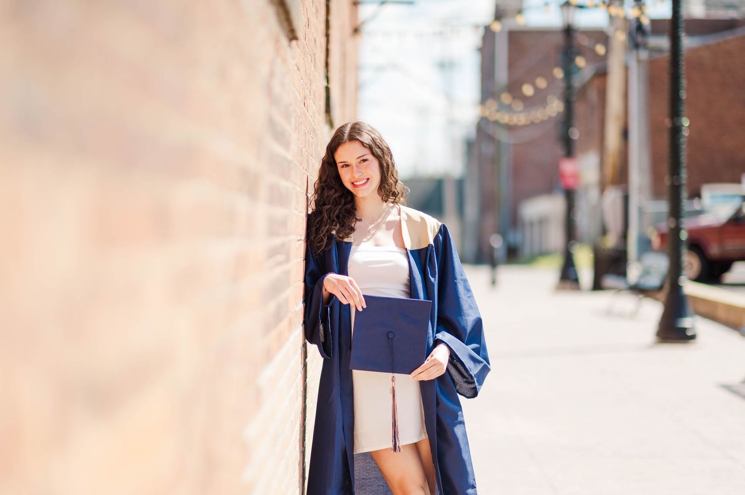 Smiling female graduate in navy blue gown leaning against brick wall holding diploma outdoors.