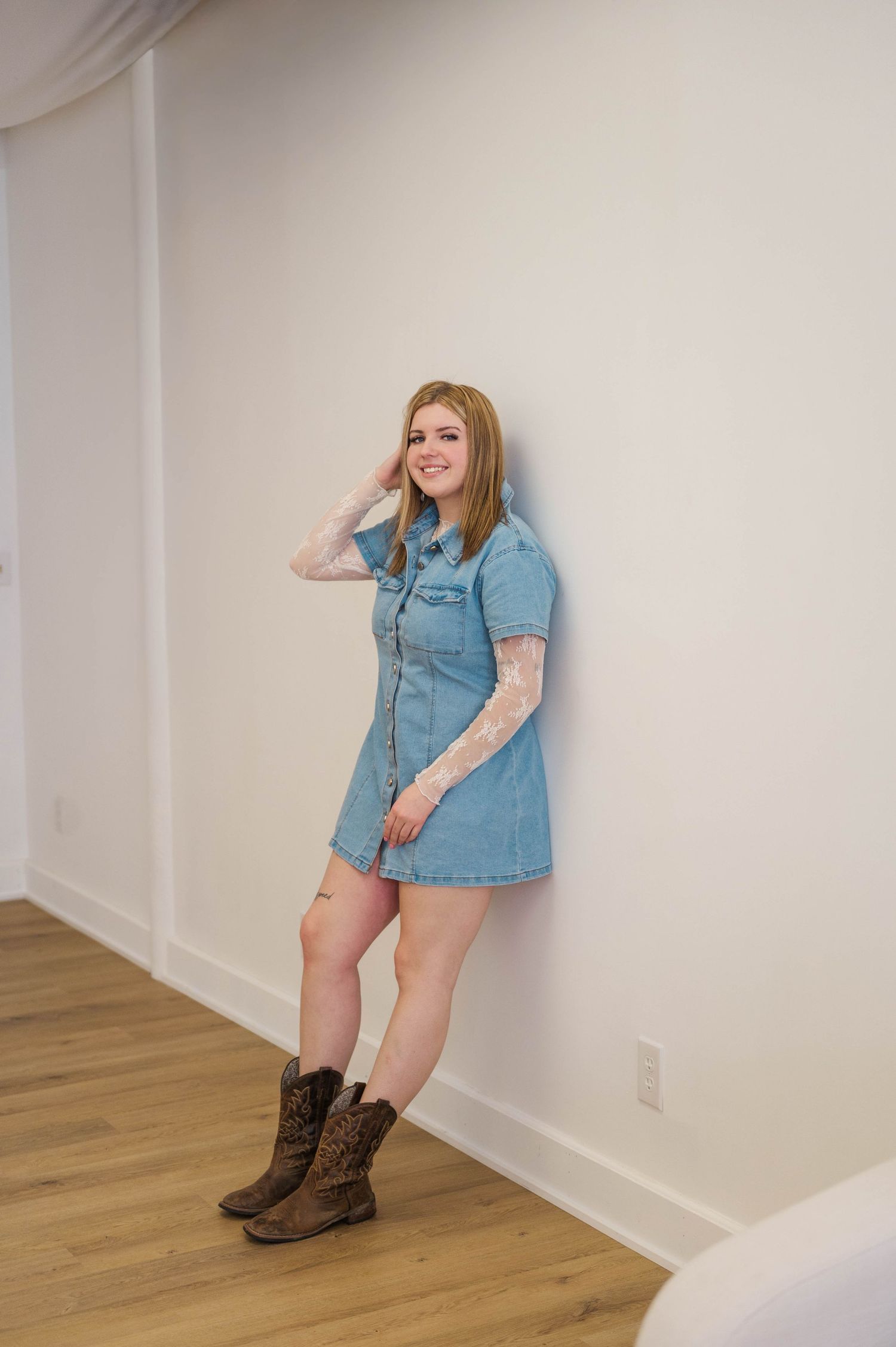 Smiling blonde woman in denim mini dress and brown cowboy boots leaning against white wall.