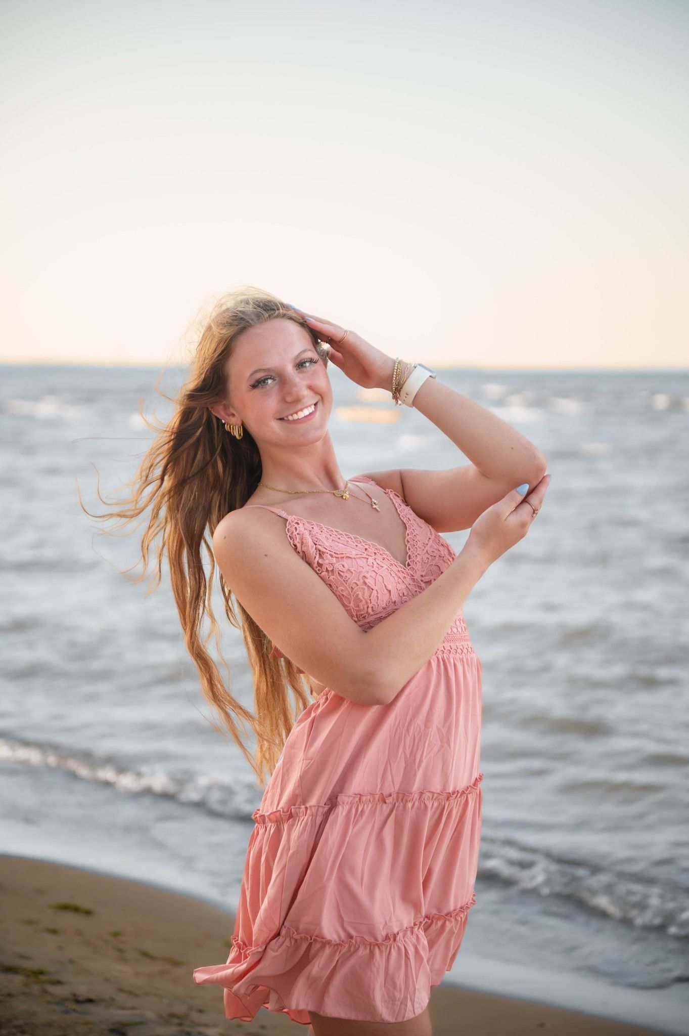 Person in pink dress posing playfully at the beach during sunset.