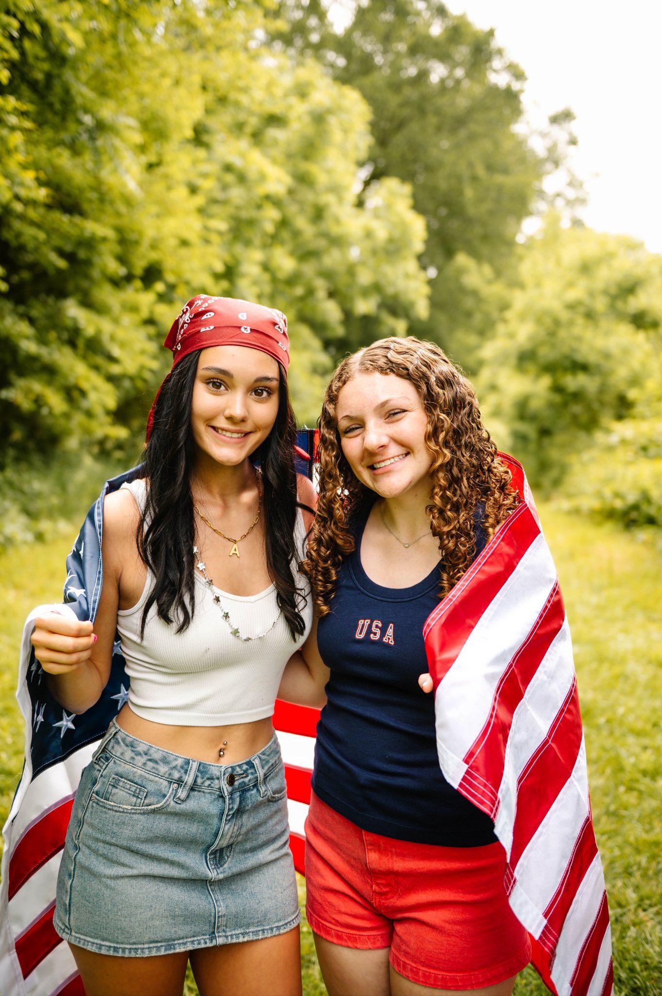 Friends in patriotic summer outfits pose with American flags against green foliage.