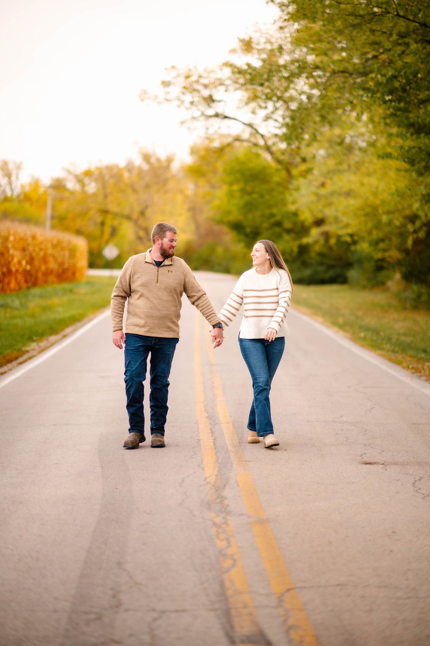 Couple walking together down empty rural road during golden hour with fall trees lining the path.