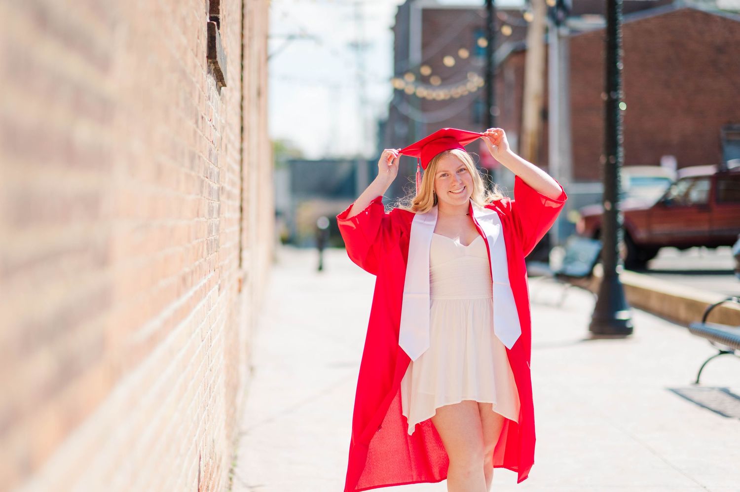 Smiling graduate in red cap and gown walking along sunny urban sidewalk adjusting her cap.