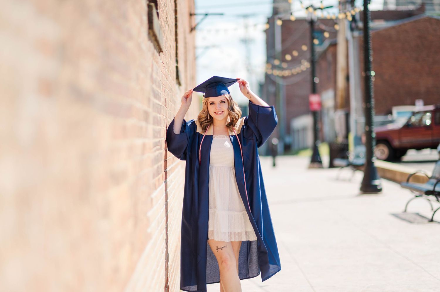 Smiling graduate in navy cap and gown leans against a brick wall on a sunny urban street.
