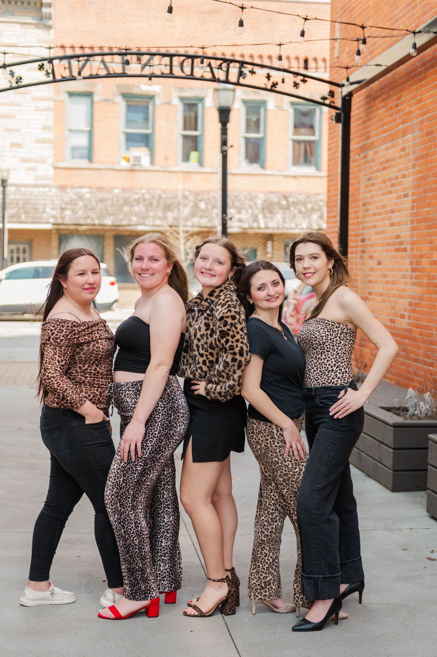 Five young women in leopard print and black outfits pose together smiling under a decorative arch.