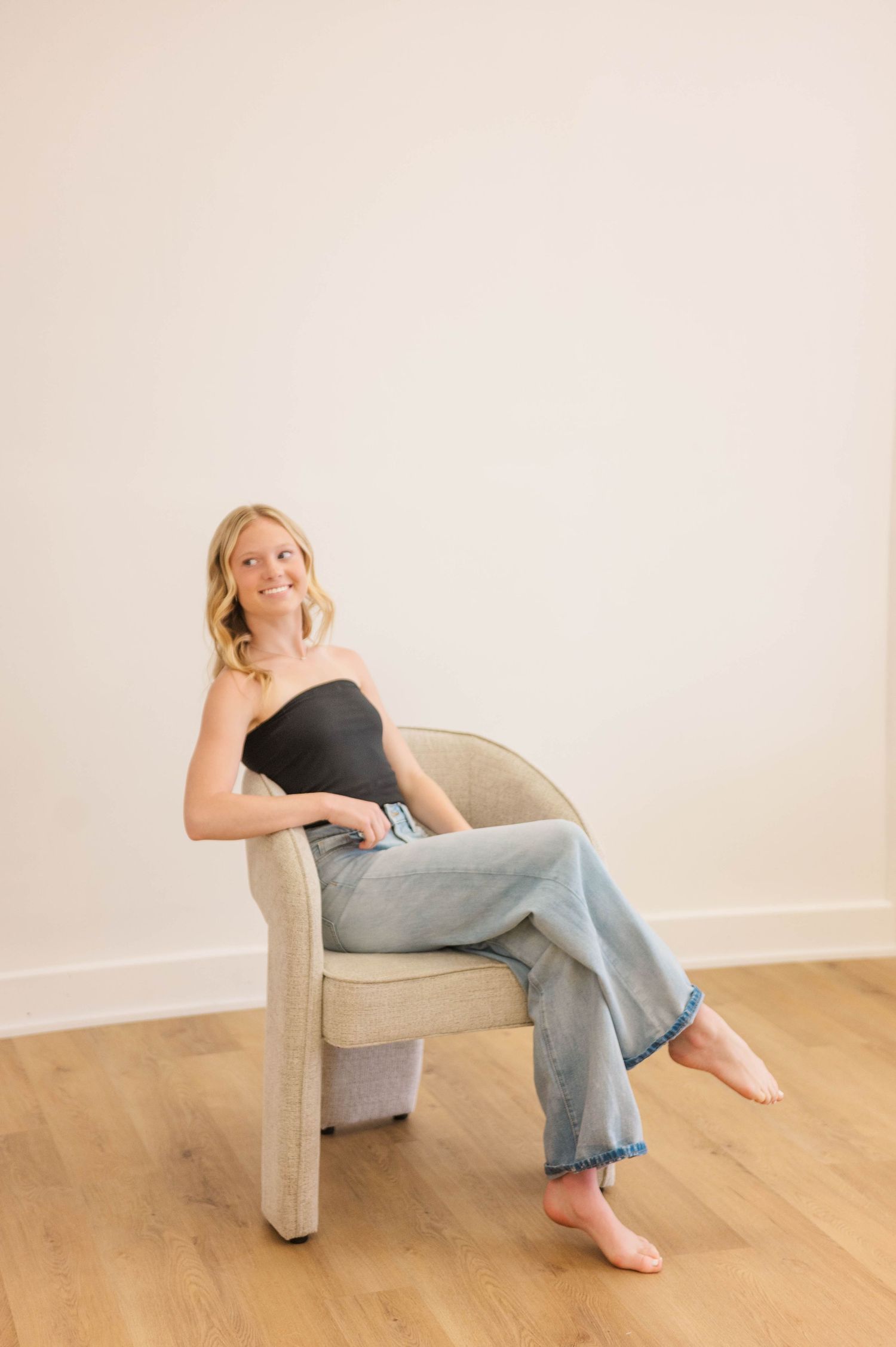 Blonde woman in black tube top and wide-leg jeans sitting barefoot in beige chair against white wall.