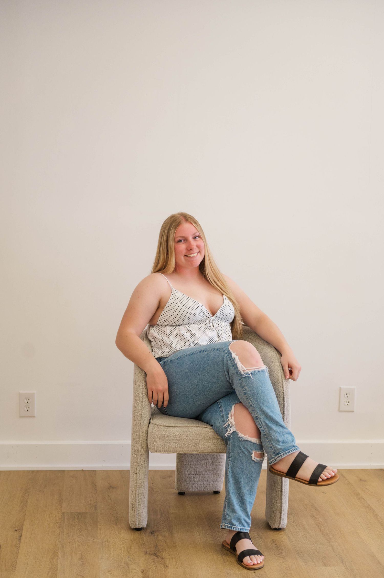 Smiling blonde woman in striped cami and ripped jeans sitting casually in a modern chair on hardwood floor.