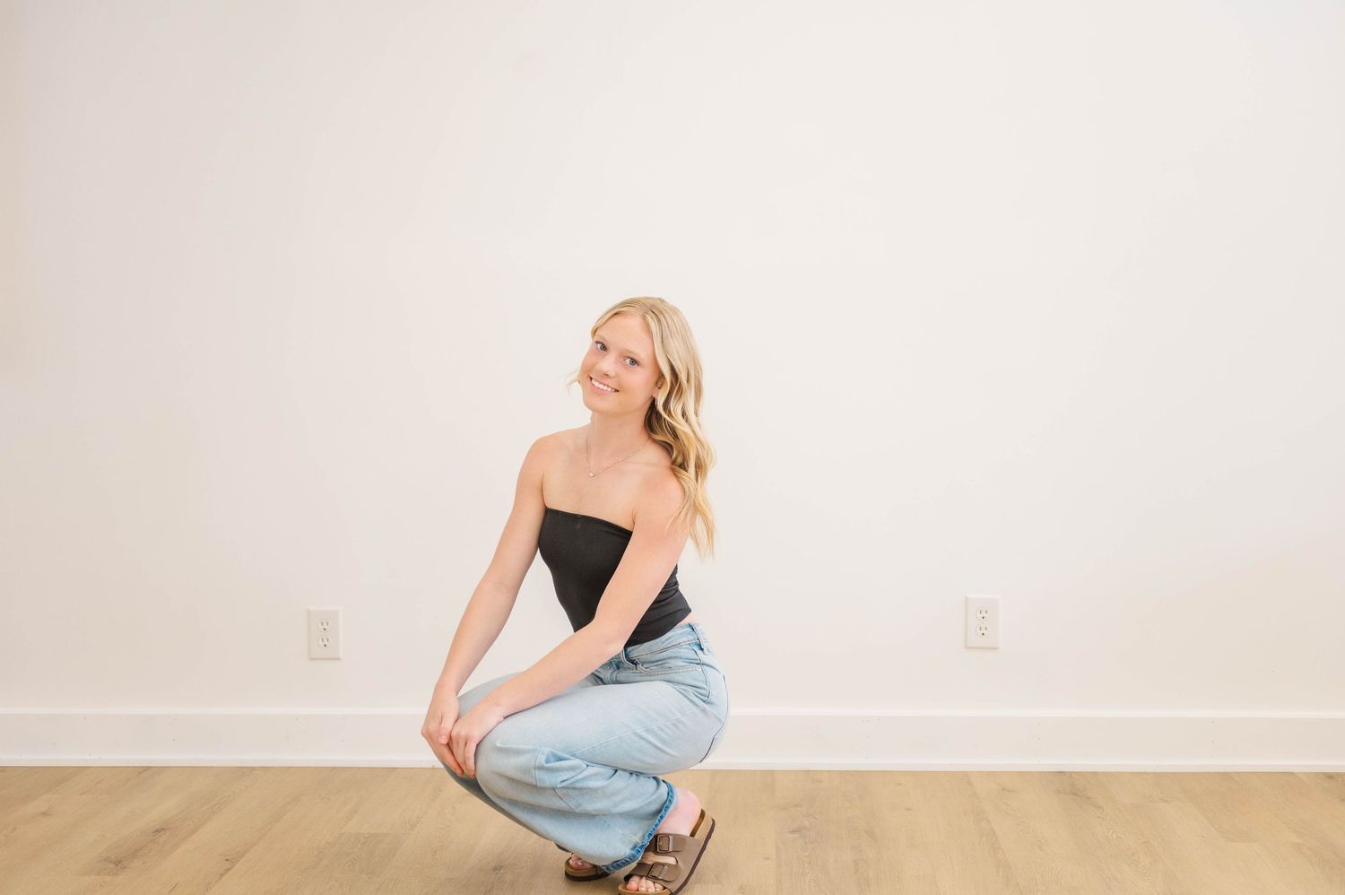 Blonde woman in black tube top and light jeans crouching on wood floor smiling over shoulder.