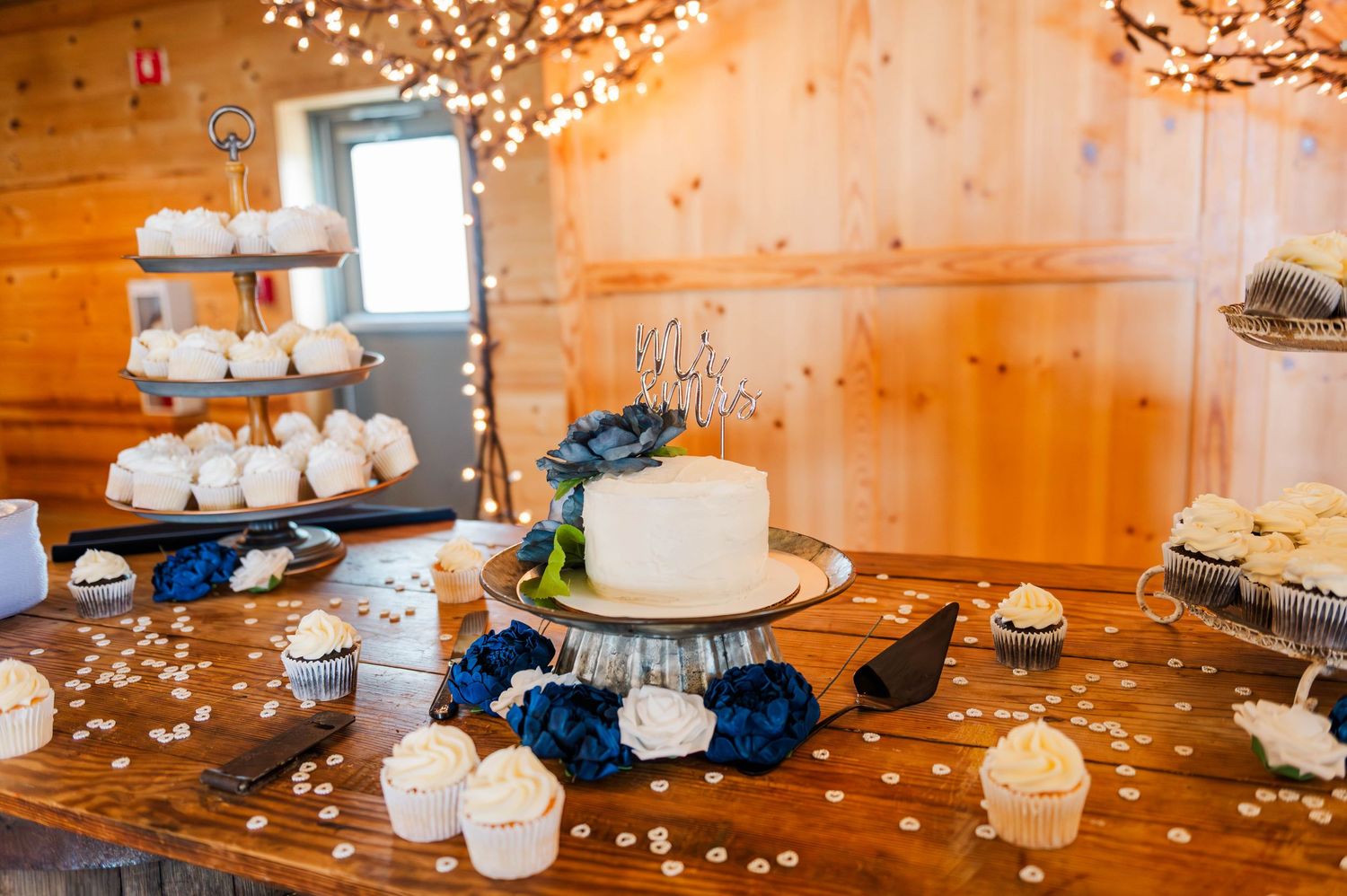 Elegant white wedding cake with navy blue accents and string lights displayed on rustic wooden dessert table.