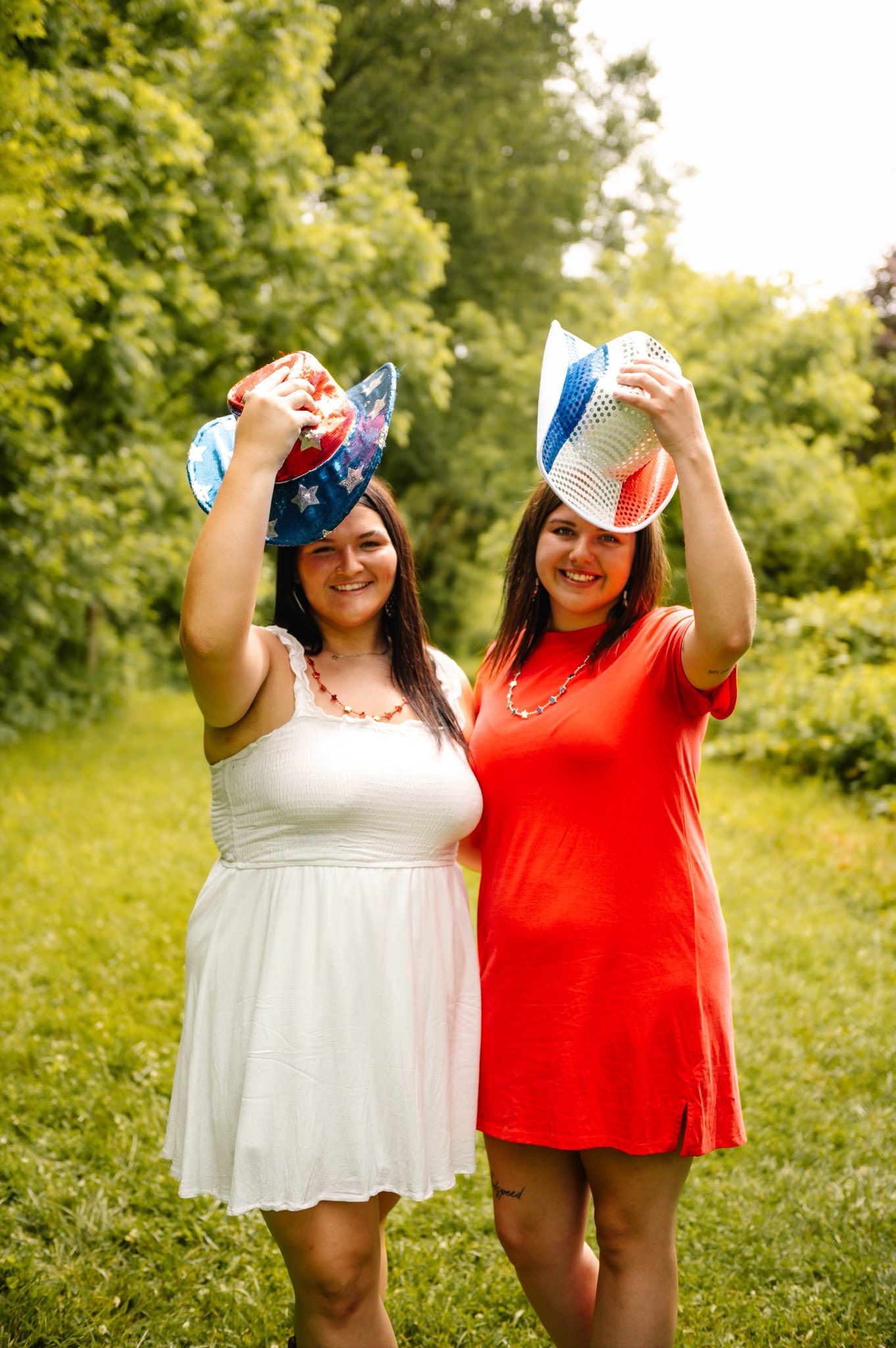 Two friends wearing patriotic colors pose playfully with baseball caps in a summer field.