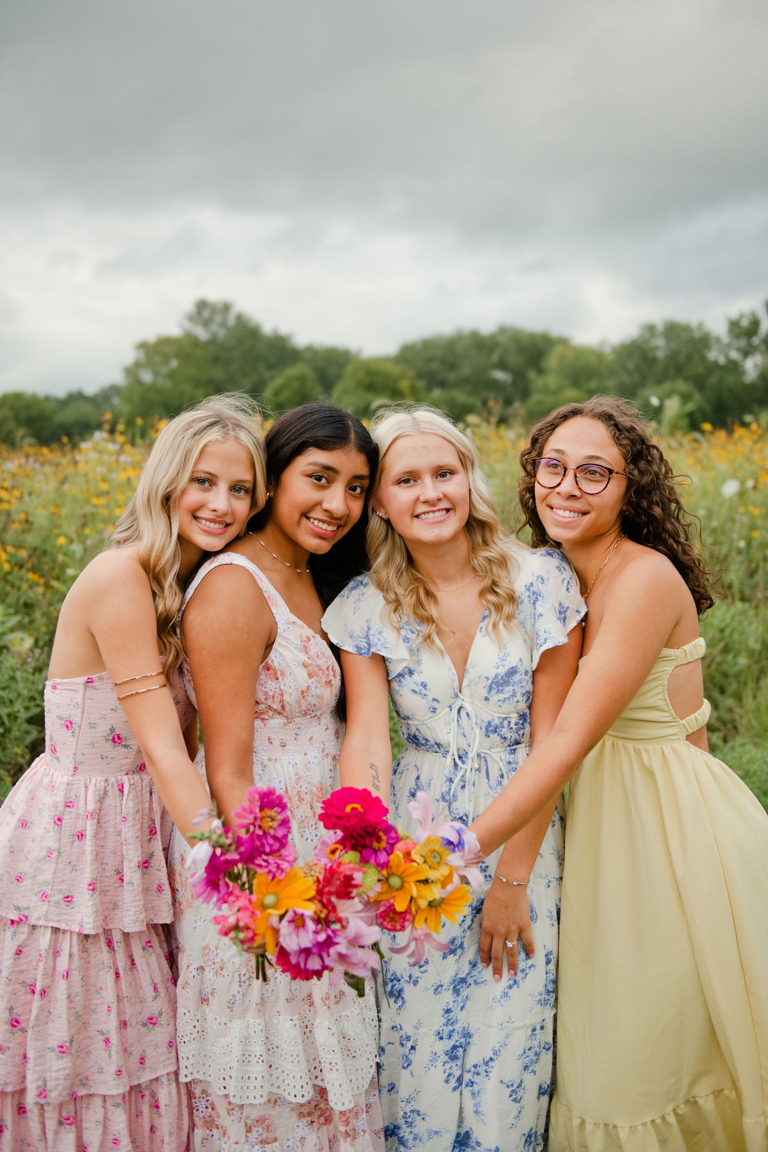 Four young women in colorful floral dresses posing with a bright bouquet in a flower field.