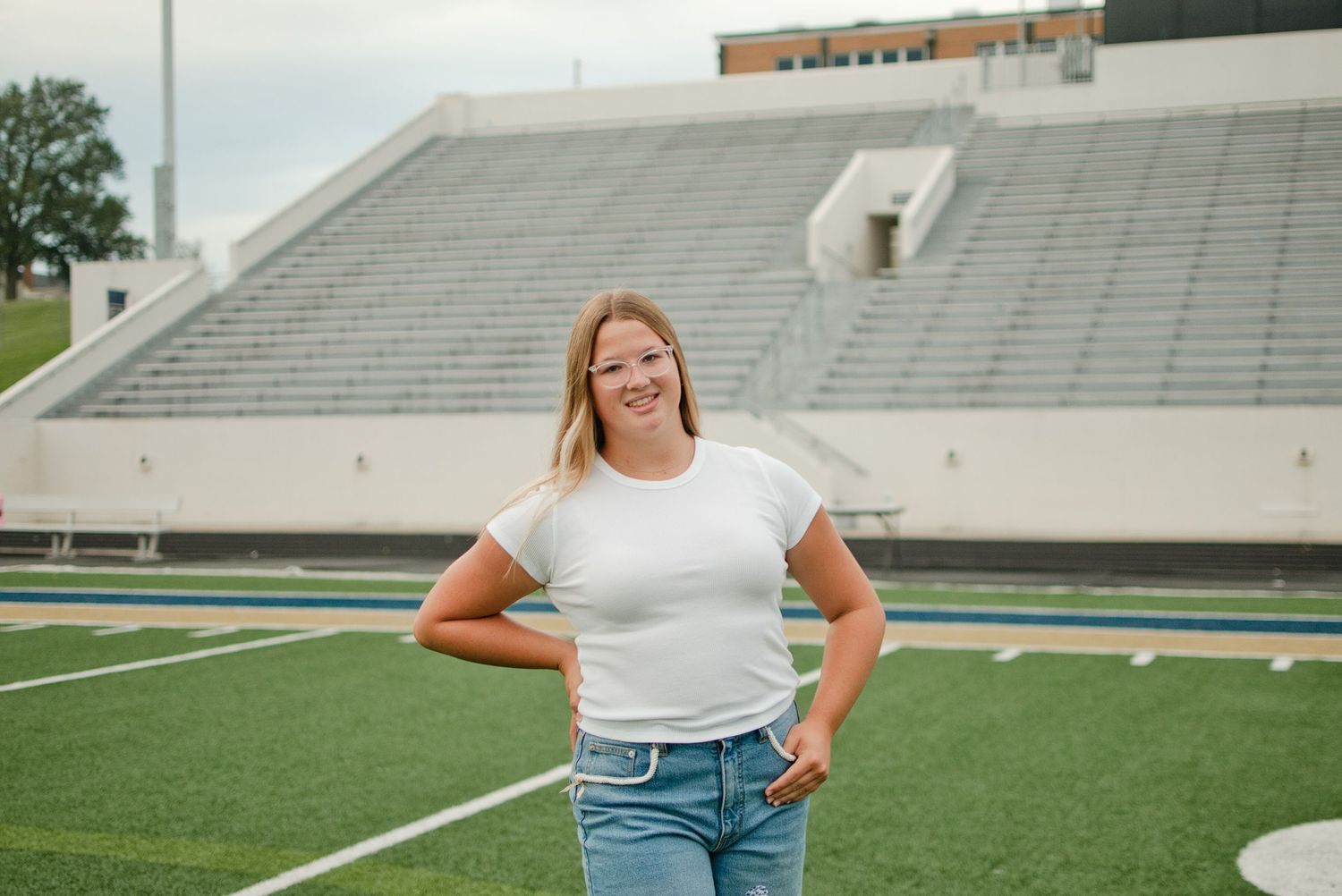 Young blonde woman smiling in white t-shirt and jeans standing on green turf football field with stadium bleachers.