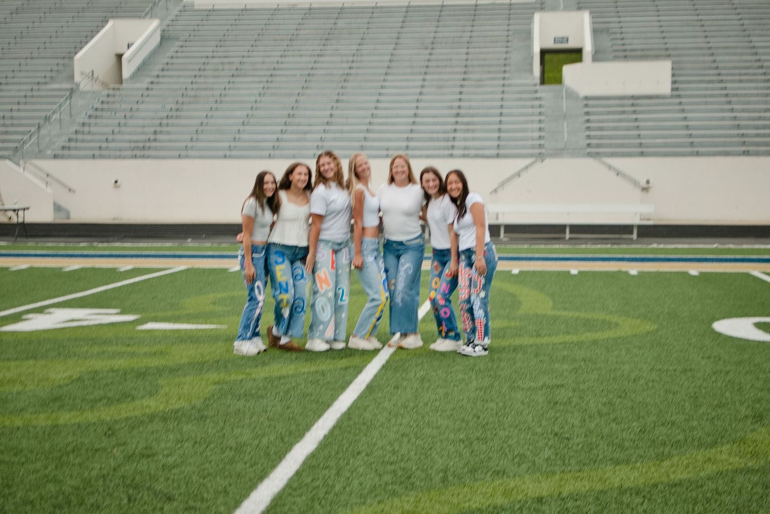 Group of seven girls in white tops and jeans posing together on a football field.