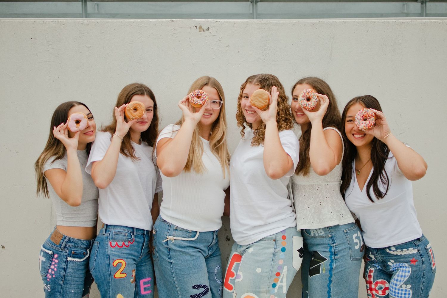 Six girls in white tops and painted jeans holding donuts over their eyes against a wall.